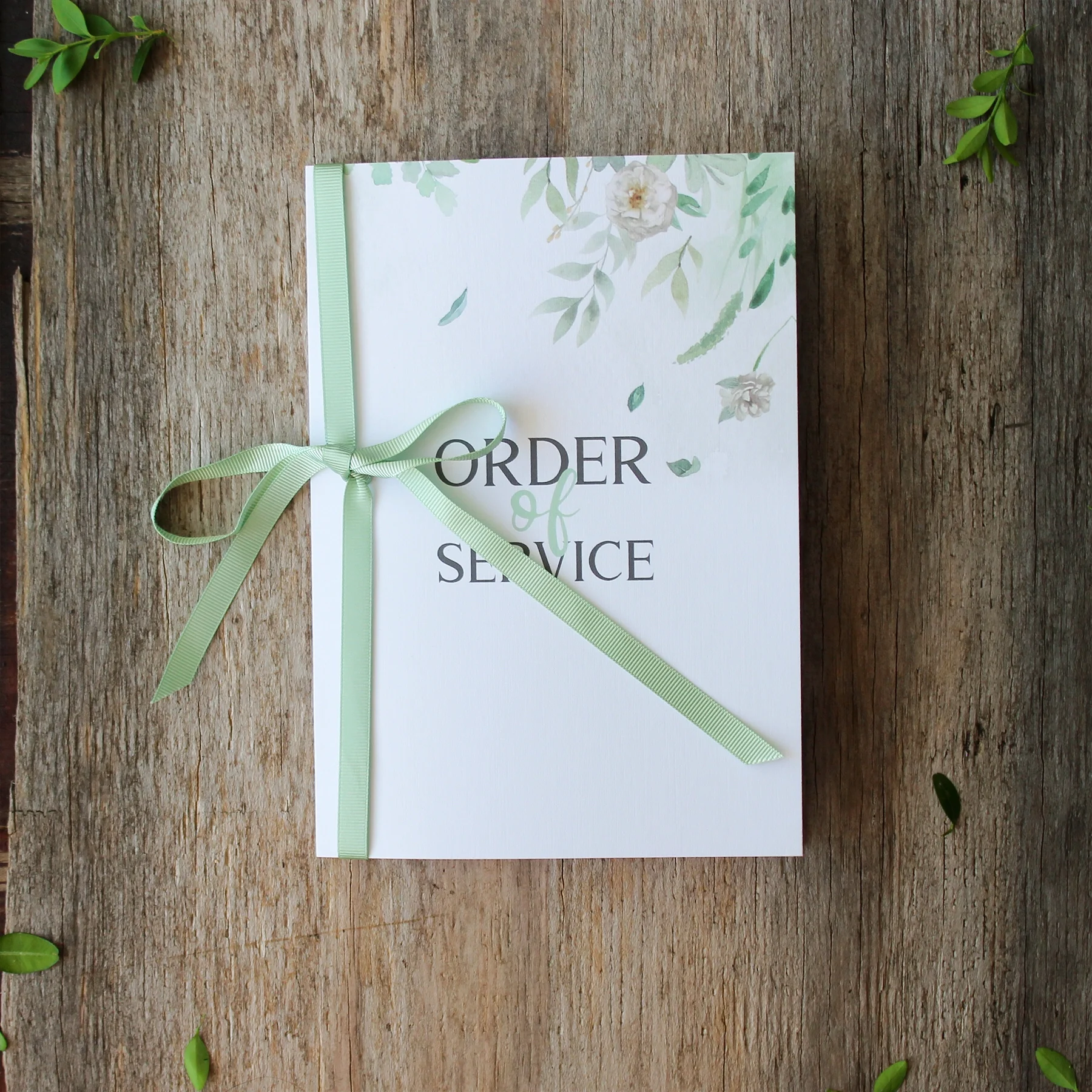 A white card with floral design and the words 'Order of Service' in the center, tied with a light green ribbon, placed on a wooden surface with small green leaves around.