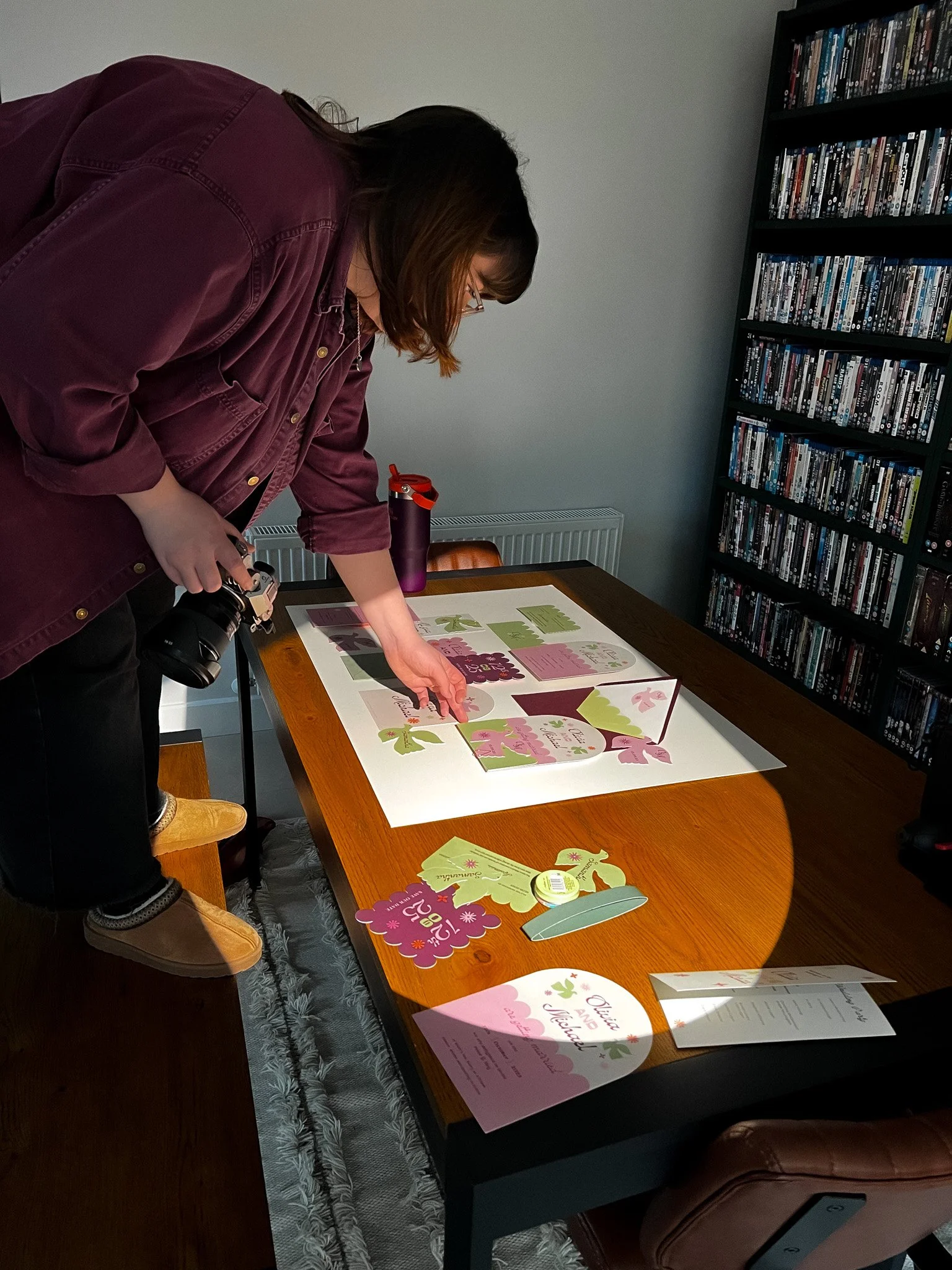 Woman arranging pink and green greeting cards on a wooden table, with a camera hanging around her neck and DVD shelves in the background.