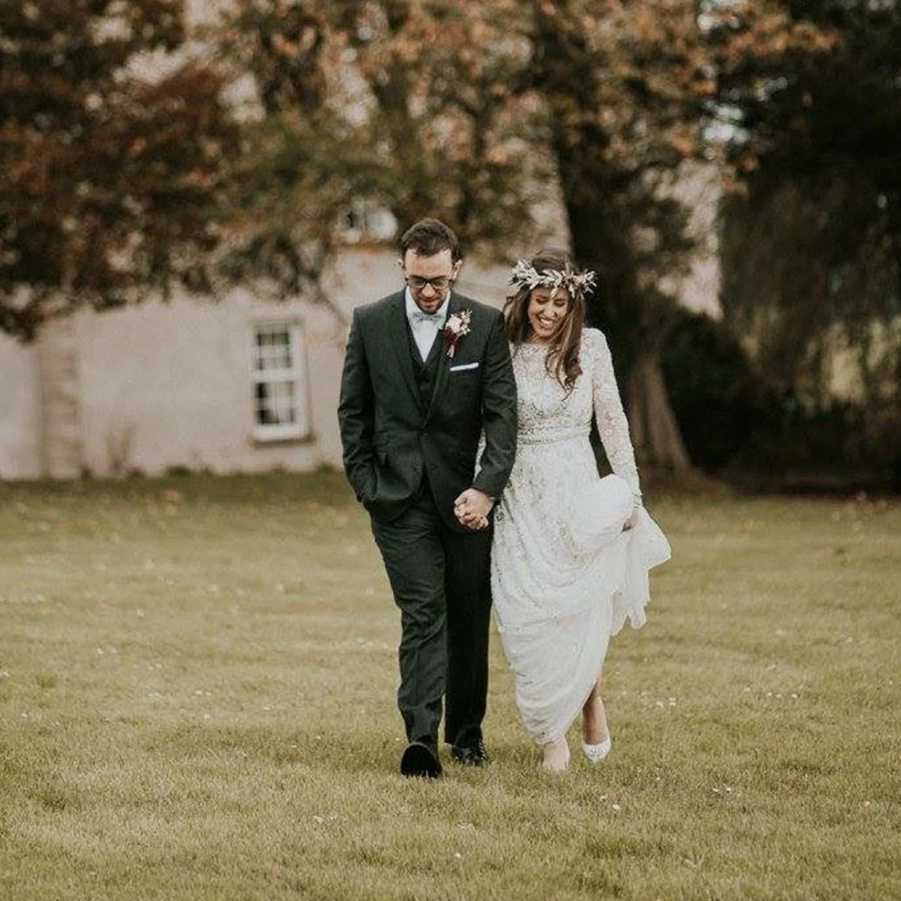 A bride and groom walking hand in hand outside on a grassy field, smiling, with trees and a building in the background. The bride is wearing a white lace wedding dress and a flower crown, while the groom is in a dark suit with glasses.