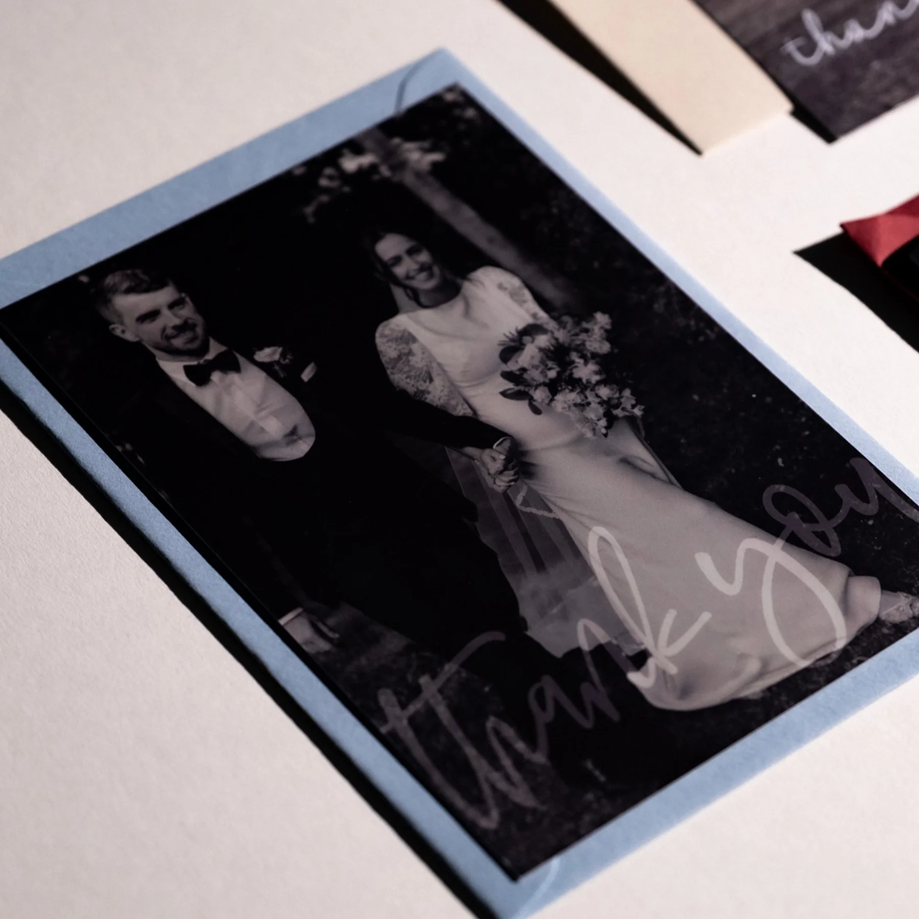 A black and white wedding photo of a bride and groom holding hands, with the words 'thank you' written on the bottom, placed on a white surface with some colorful cards nearby.