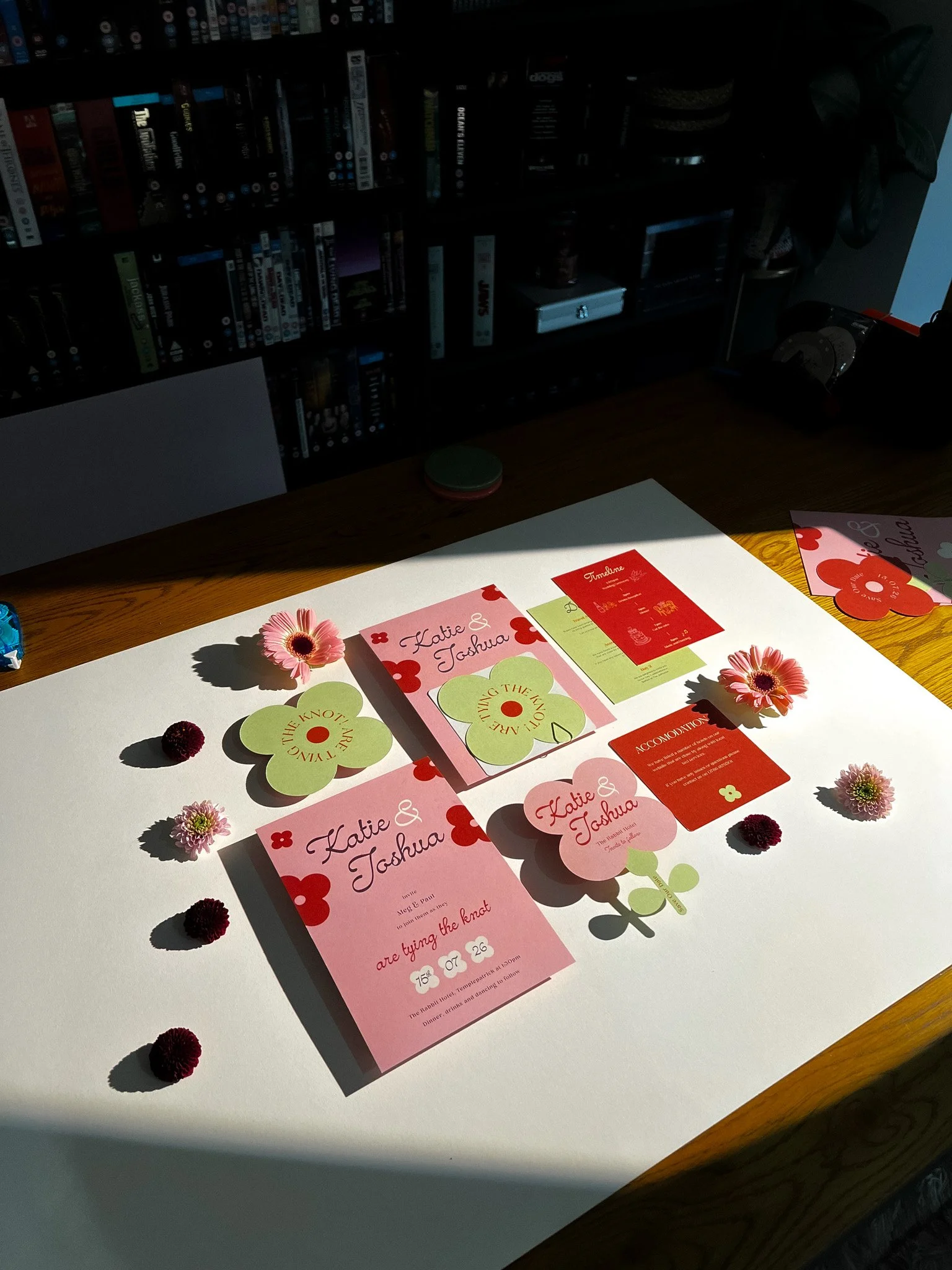 Wedding invitation suite with pink and red floral themed stationery, surrounded by small flowers on a white surface.