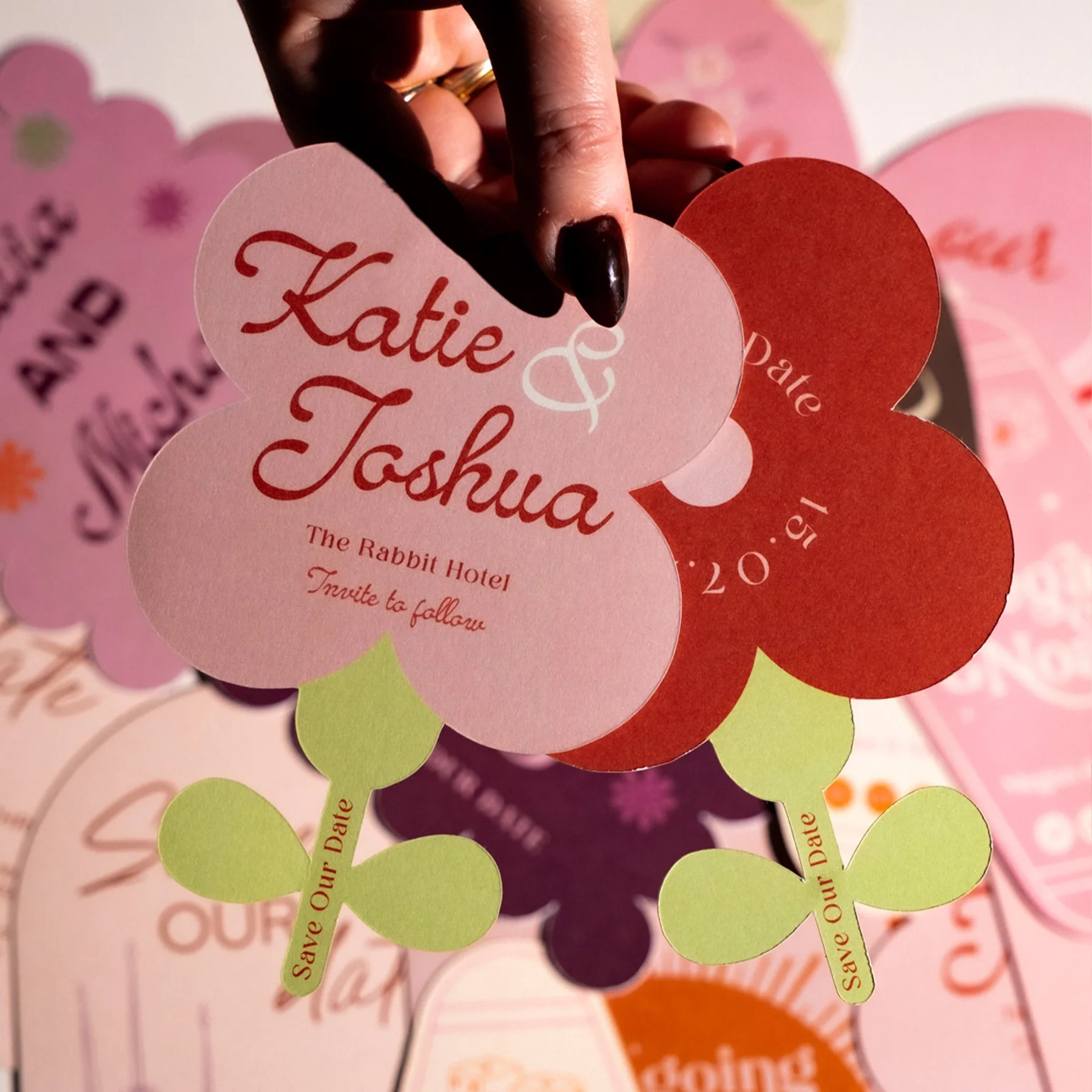 A wedding invitation shaped like a flower with pink, red, purple, and yellow petals. The center petal displays the names Katie & Joshua in red cursive font, with details of the event below. Green leaves with 'Save Our Date' written on them extend fro