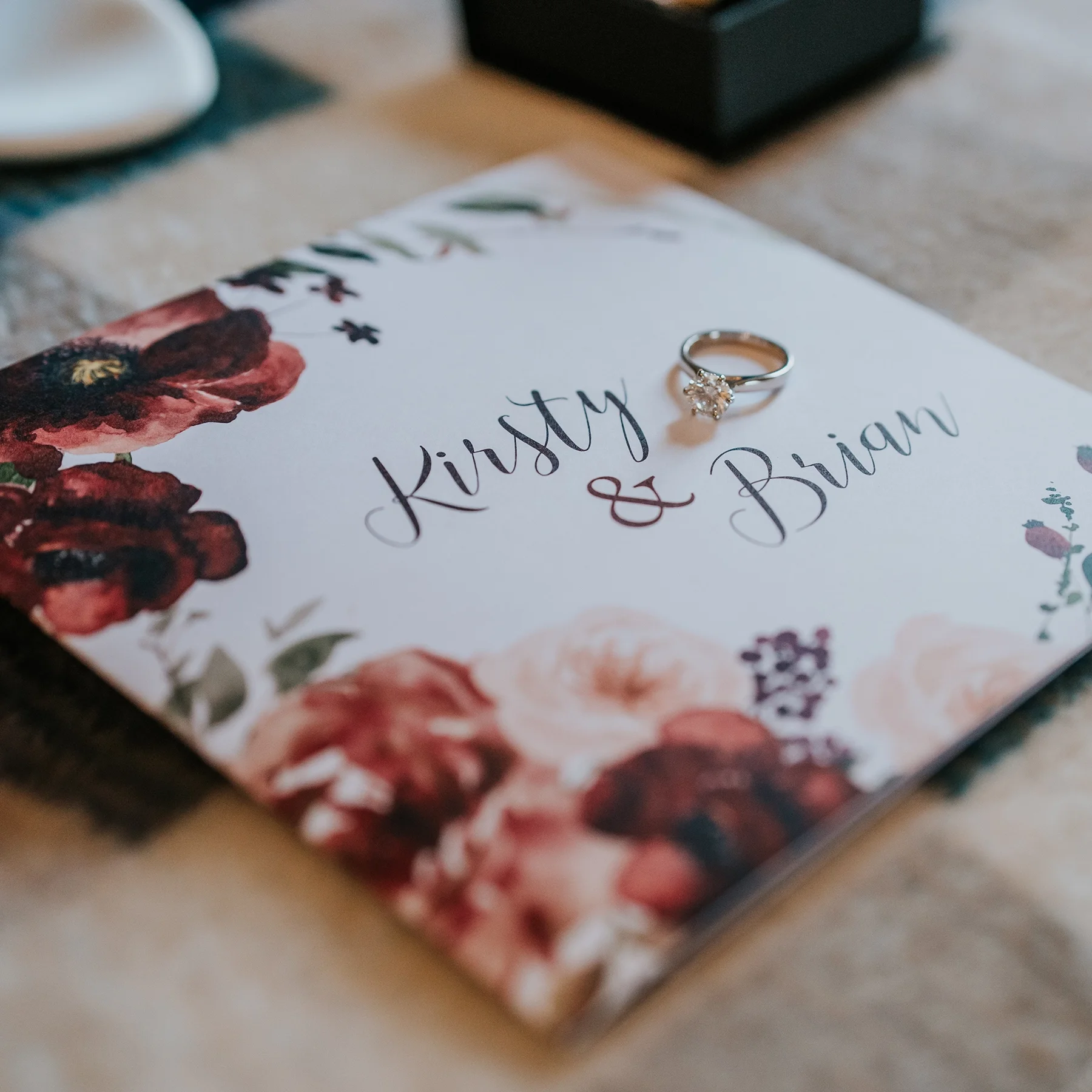 A wedding invitation with floral designs and the names "Kirsty & Brian" written in elegant script. A diamond engagement ring is placed on top of the invitation.