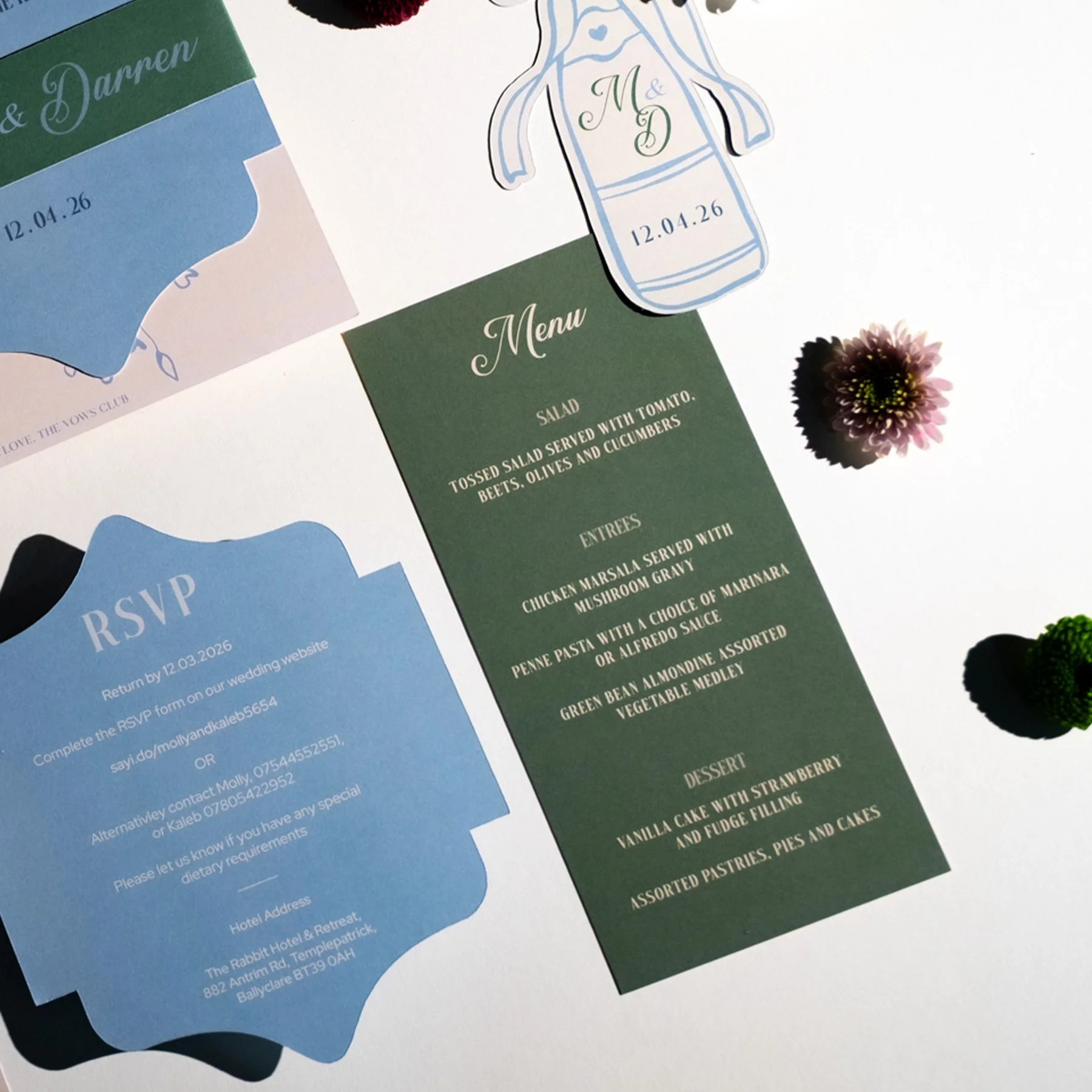 Wedding invitation with a green menu card, a blue RSVP card, and a small gift tag with the initials M & D and the date 12.04.26. There are also some flowers and decorative items on the table.