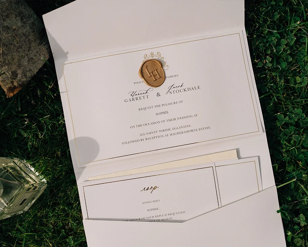 Wedding invitation with a gold wax seal on cream-colored paper, inside a white envelope, placed on grass.