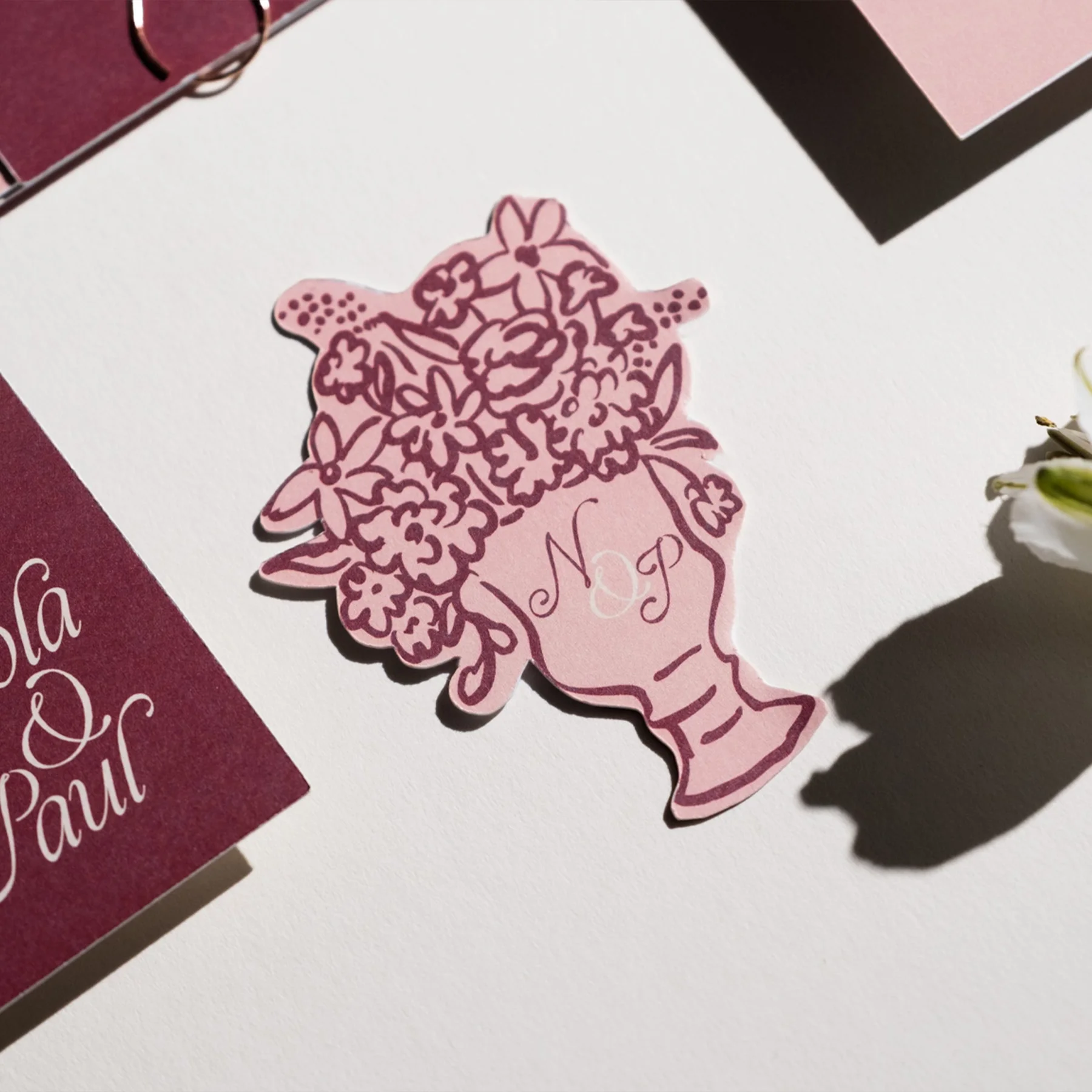 A decorative paper cutout of a woman's head with flowers as hair, with the initials 'M O P' in the middle, placed on a white surface near a burgundy card with white cursive writing.