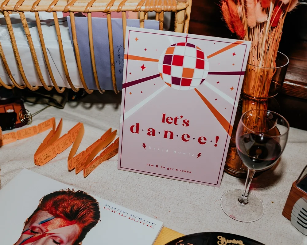 A promotional display for a dance event featuring a large card with a disco ball graphic and the text "let's dance!" by David Bowie. There are orange tickets, a wine glass, and a vase with dried flowers on the table.