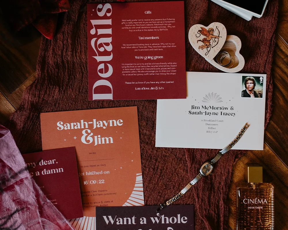 Wedding invitation suite that includes an invitation card, RSVP card, gift list, and a plane ticket-style invite with address details, along with a wristwatch, a glass perfume bottle, a small heart-shaped card with Cupid illustration, and a ring on a