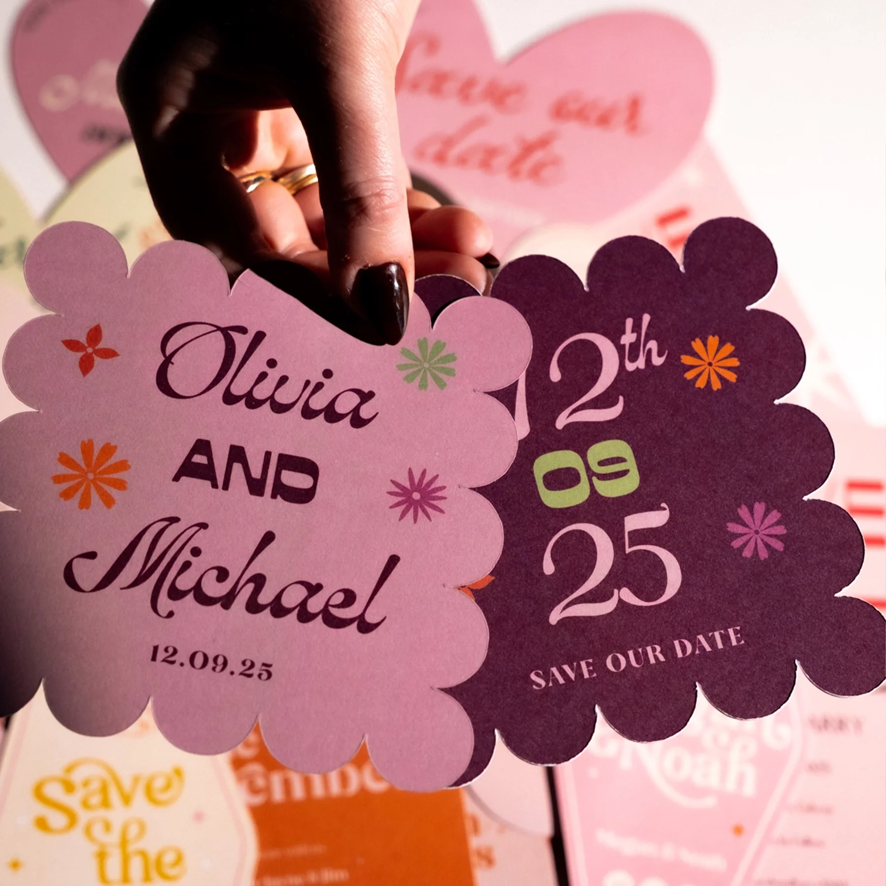 Close-up of a hand holding pink and purple floral-shaped bookmarks with wedding date and names, featuring colorful small flowers and text for a wedding save-the-date card.