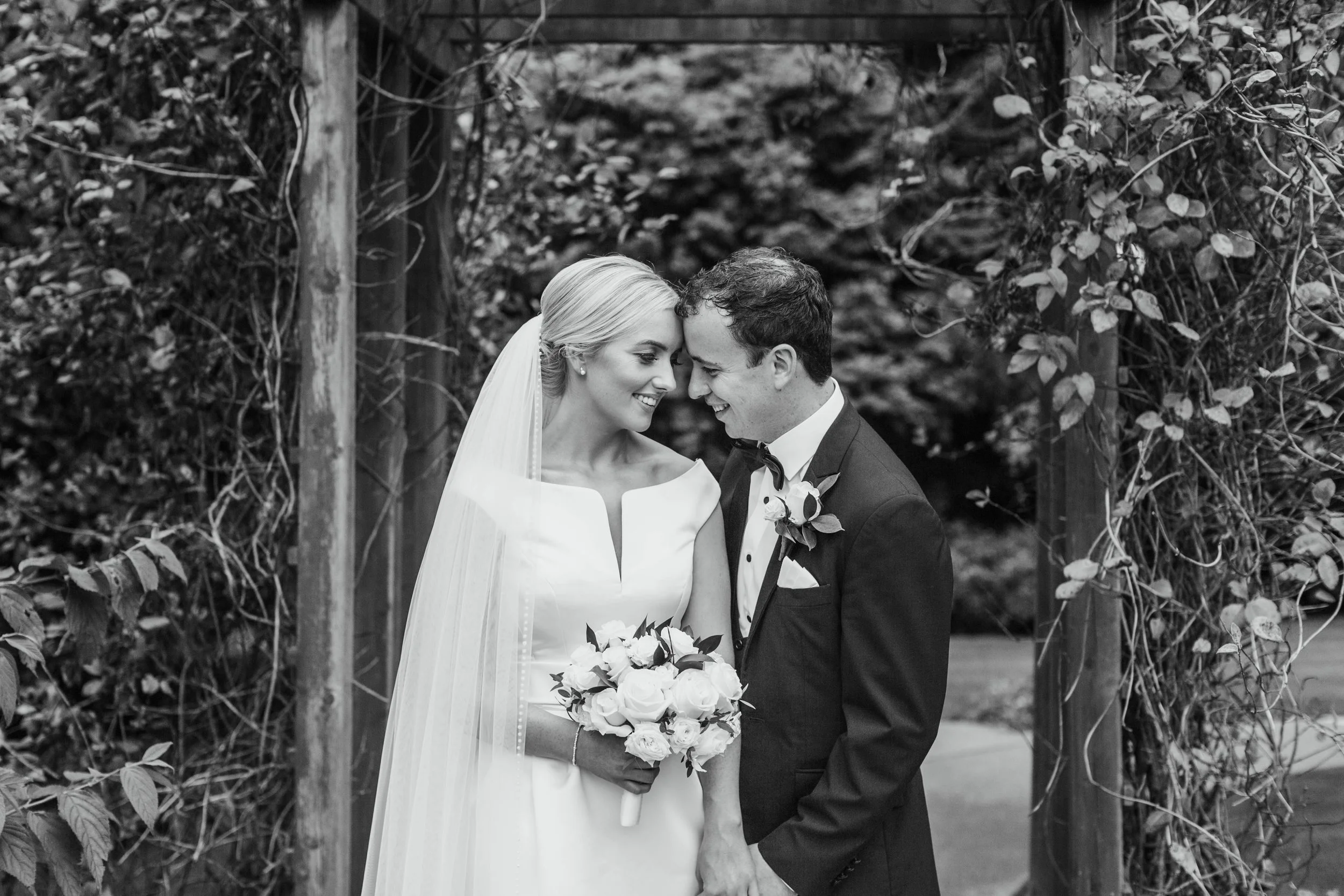 Black and white photo of a bride and groom on their wedding day, smiling and touching foreheads, with the bride holding a bouquet of flowers and surrounded by a floral arch.