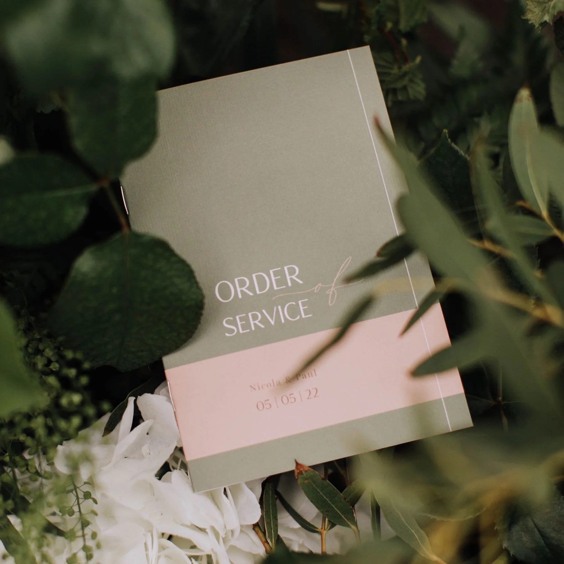 A light green and pink card titled 'Order of Service' is placed on green leaves and white flowers, with names 'Nicole & Paul' and date '05/05/22'.