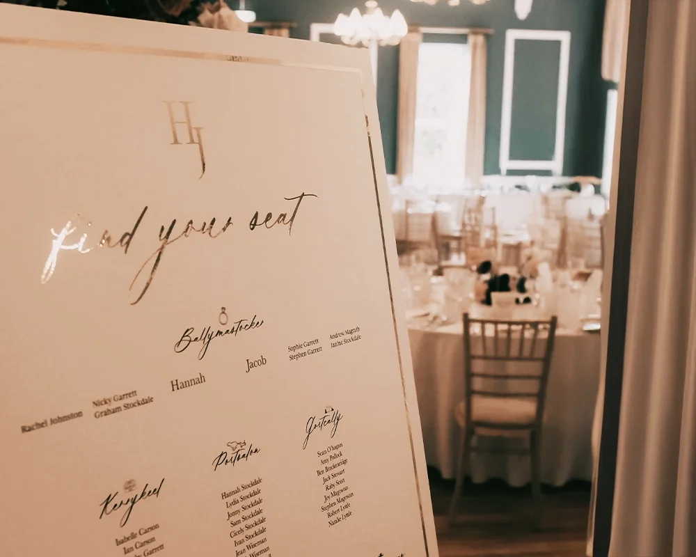 Wedding seating chart with the heading 'find your seat', listing tables named Ballymaster, Galway, Potatie, Repeval, and Gully, with guest names under each table, inside decorated wedding reception venue.