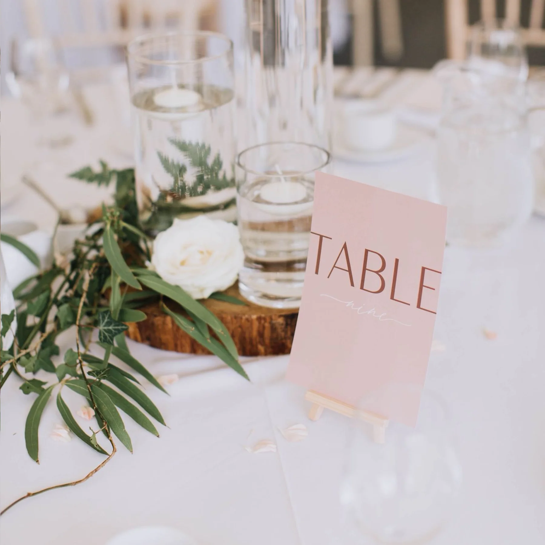 Table setting with a pink table sign and glassware, featuring greenery and a white flower on a wooden accent at a formal event.