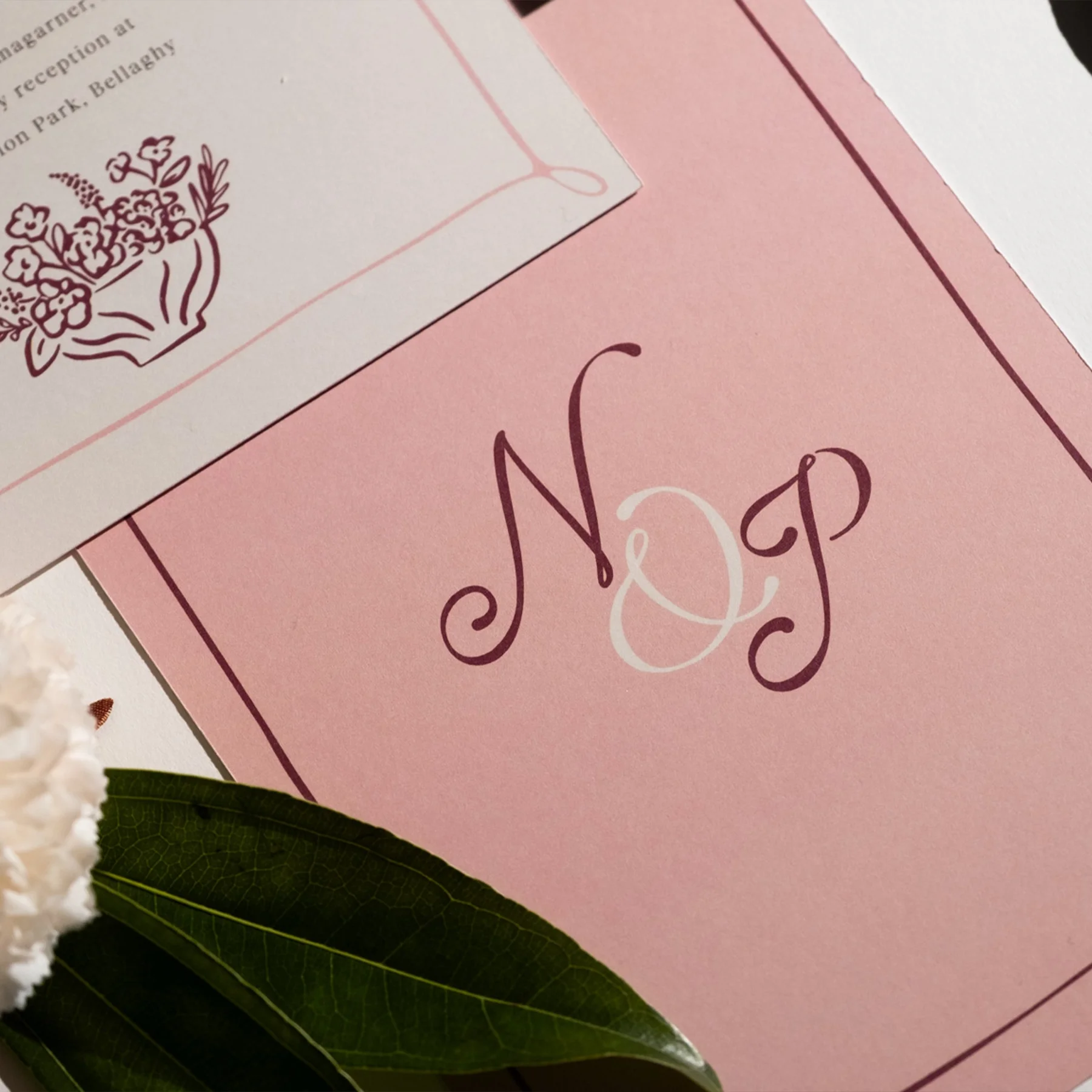 A pink card with elegant script initials 'N & P' and a white floral border, accompanied by a white flower and green leaves.
