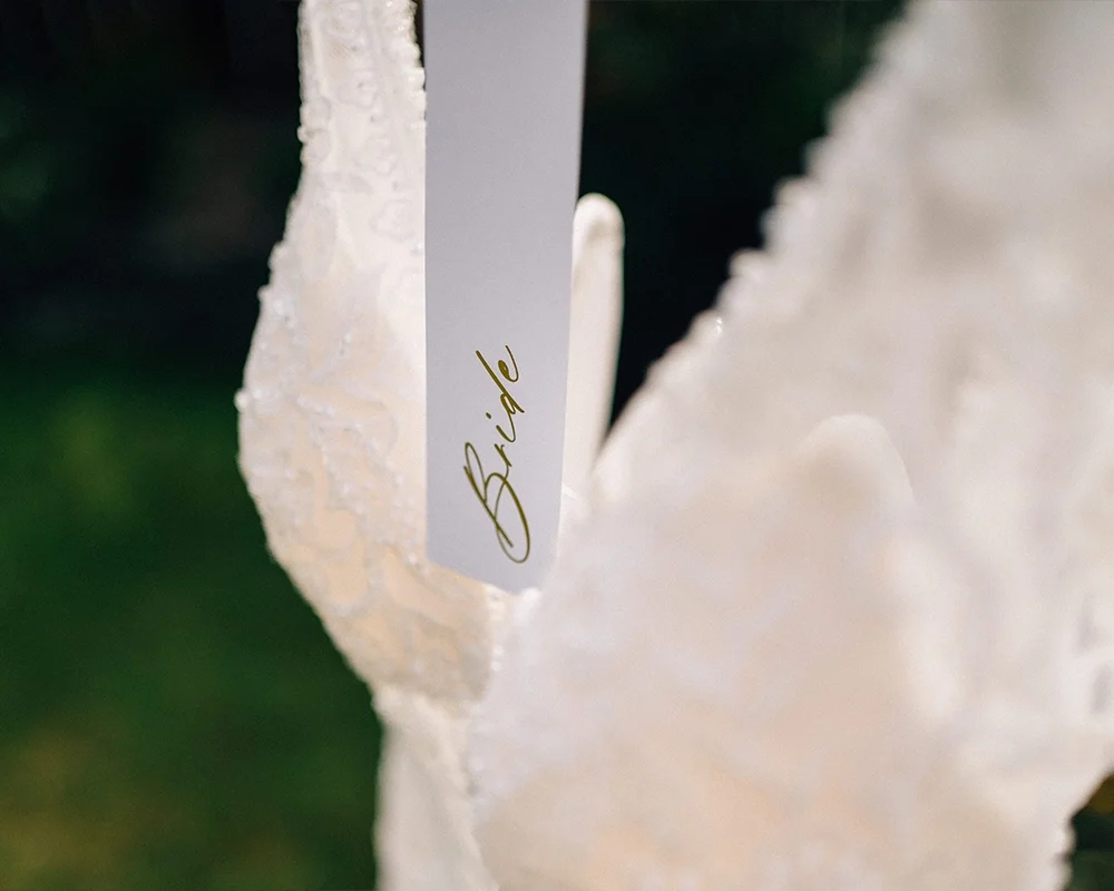 A white wedding dress with a 'Bride' tag attached, hanging on a hanger.