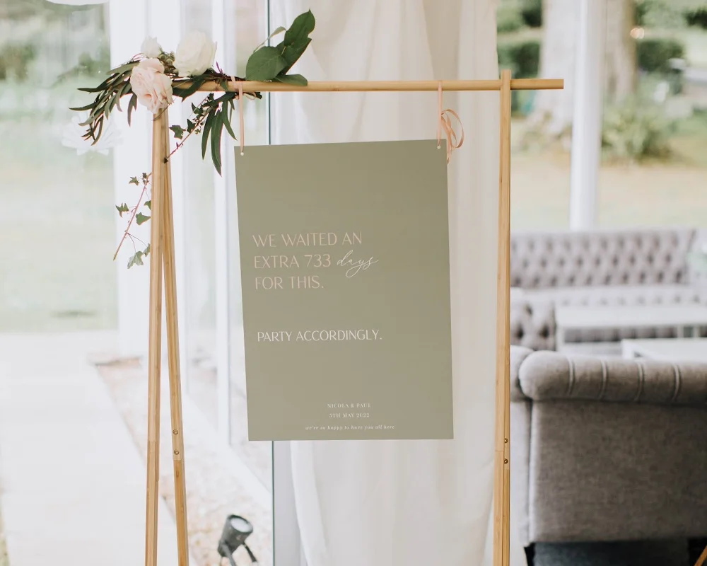 A standing sign with a floral decoration at the top left corner, displaying a message about waiting 733 days for a celebration, on a wooden frame inside a bright room with large windows and a gray couch in the background.