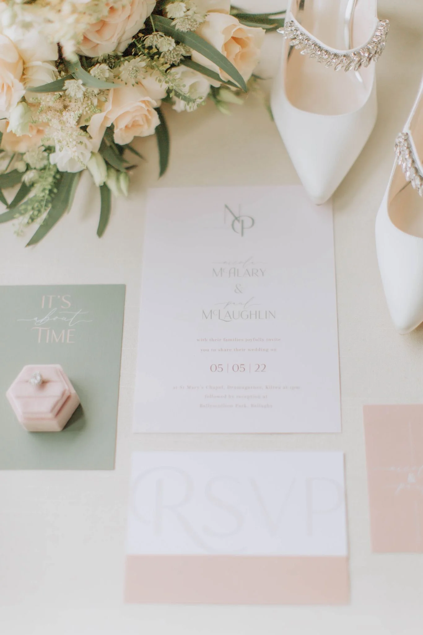Wedding invitation card with monogram, floral bouquet, pair of white high-heeled shoes with rhinestone embellishments, and a pink ring box.