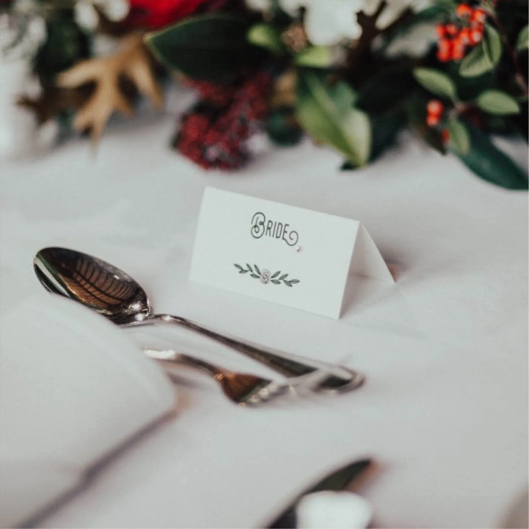 A silver spoon on a white table with a paper tent card labeled 'BRIDE' in a decorative font, with greenery and red berries in the background.