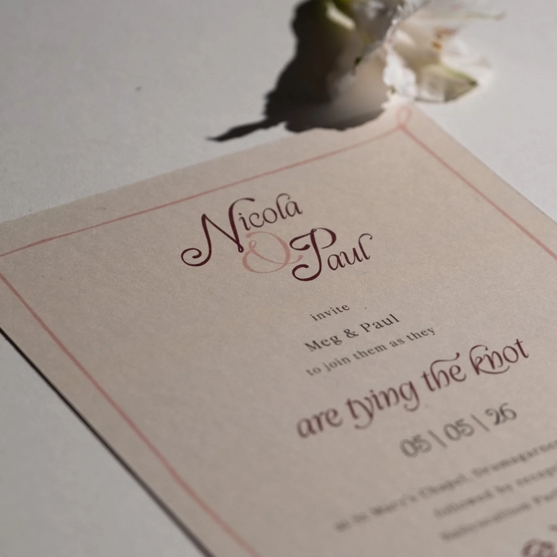 Close-up of a wedding invitation with elegant cursive text, including the names Nicola and Paul, and a small white flower on the top left corner.