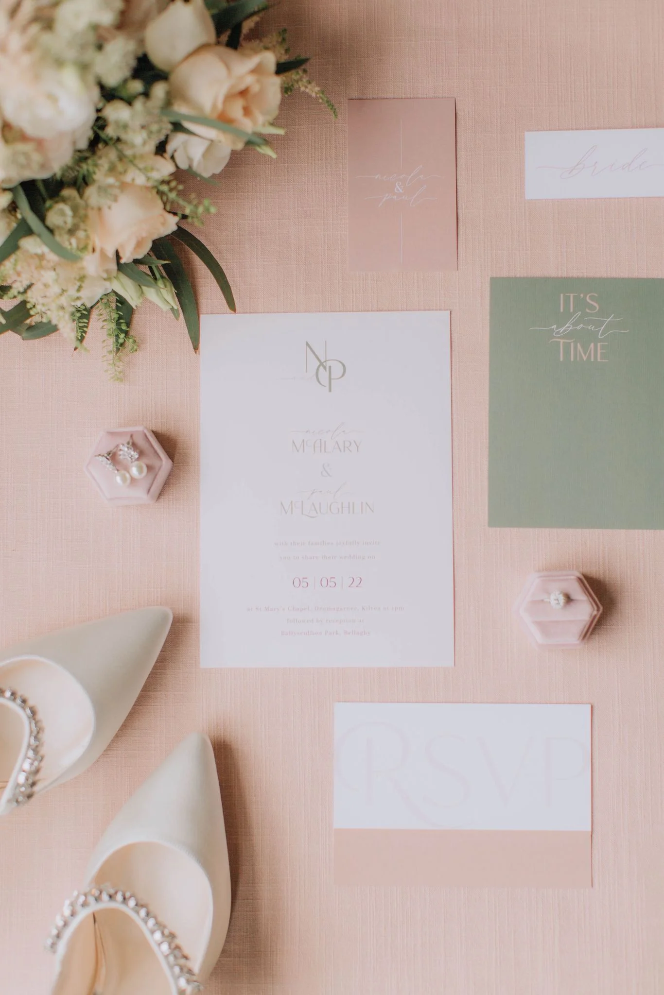 A wedding invitation display with a floral bouquet, white shoes with embellishments, and various stationery including a 'RSVP' card, a green card that says 'It's about time,' a pink card, and a white invitation card with details of the wedding of McA