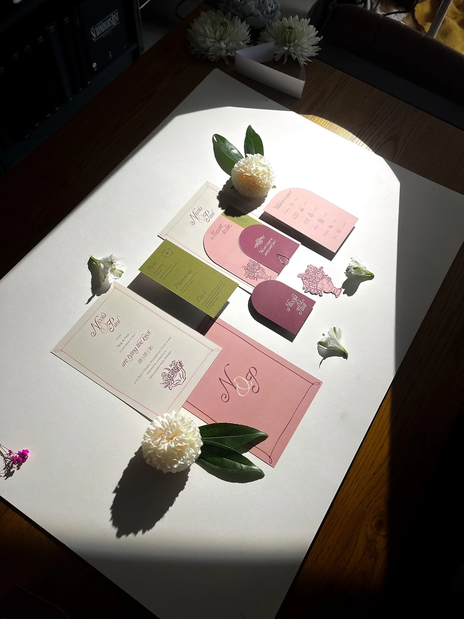 Wedding invitation display with floral decorations on a table under natural sunlight. The invitation features pink and green cards, along with white flowers and green leaves arranged around it.