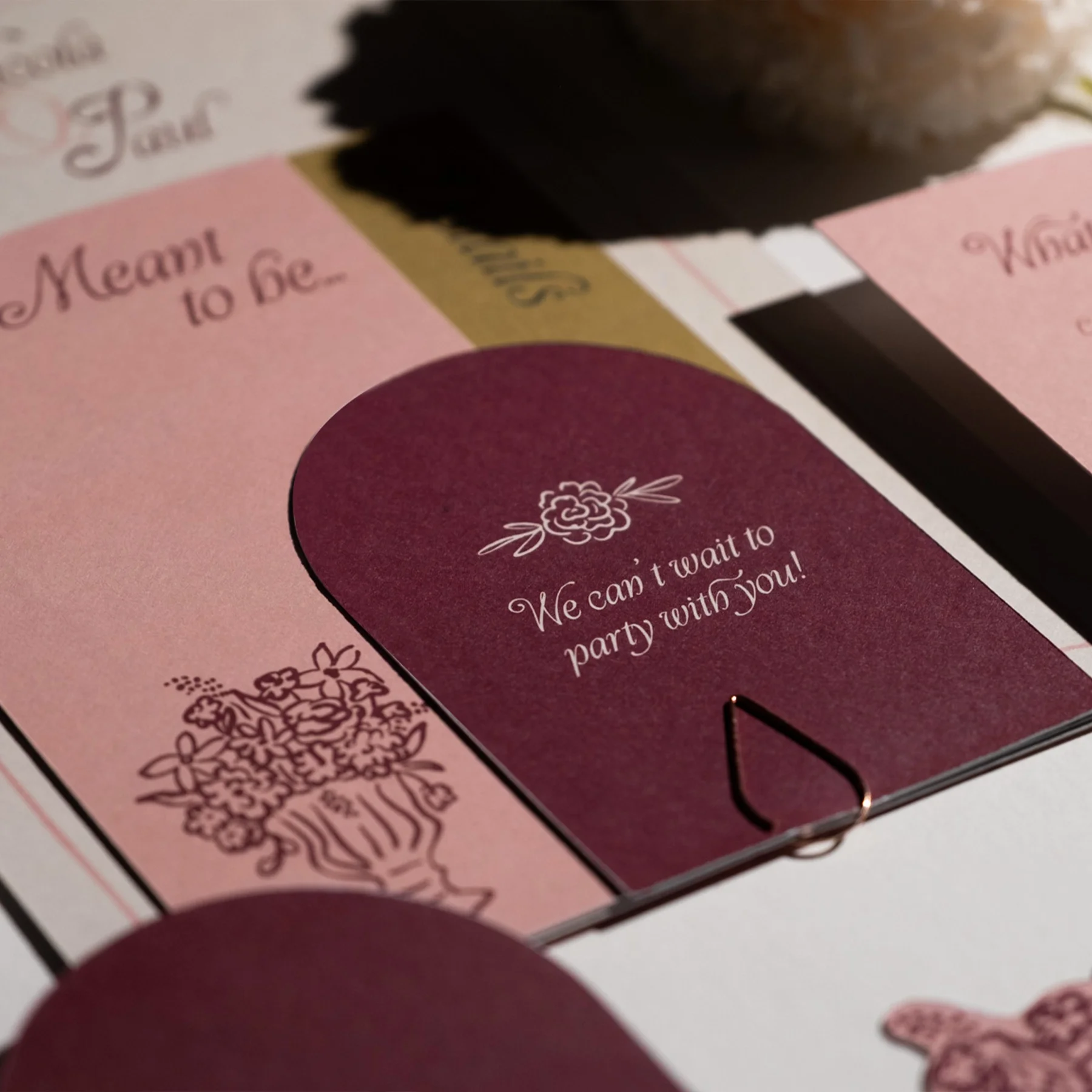 Close-up of a pink and purple card with the text "We can't wait to party with you!" and floral illustrations, part of a wedding or party invitation.