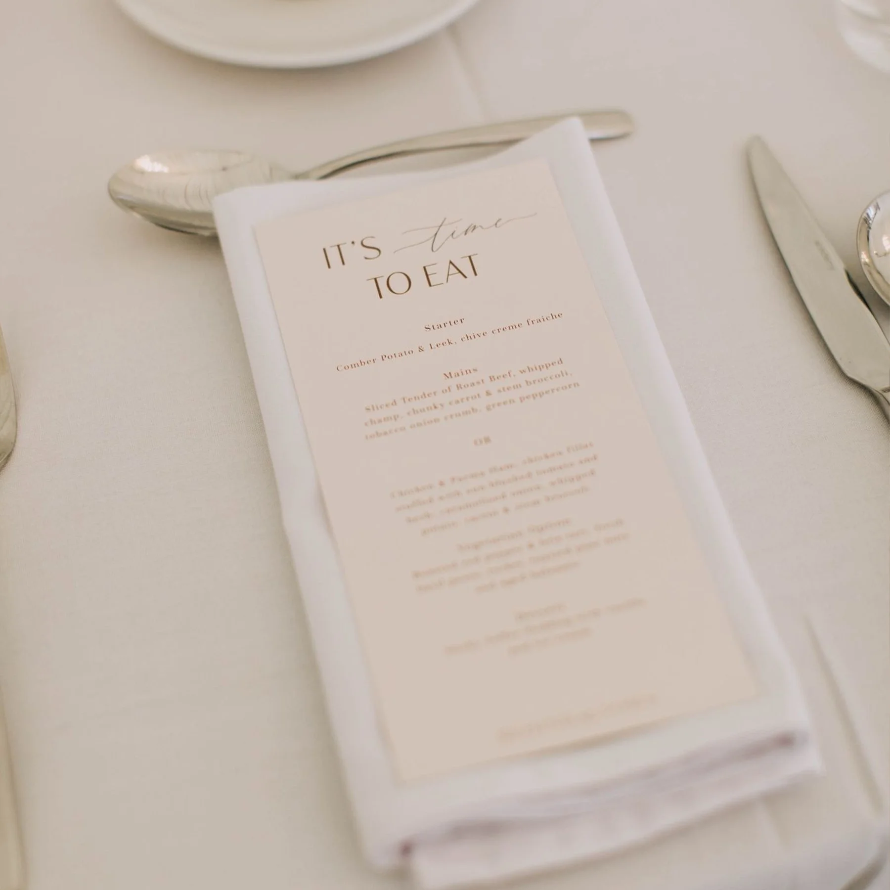 Elegant table setting with a printed menu titled 'It's time to eat' on a cloth napkin, surrounded by silverware with a knife and spoon visible.