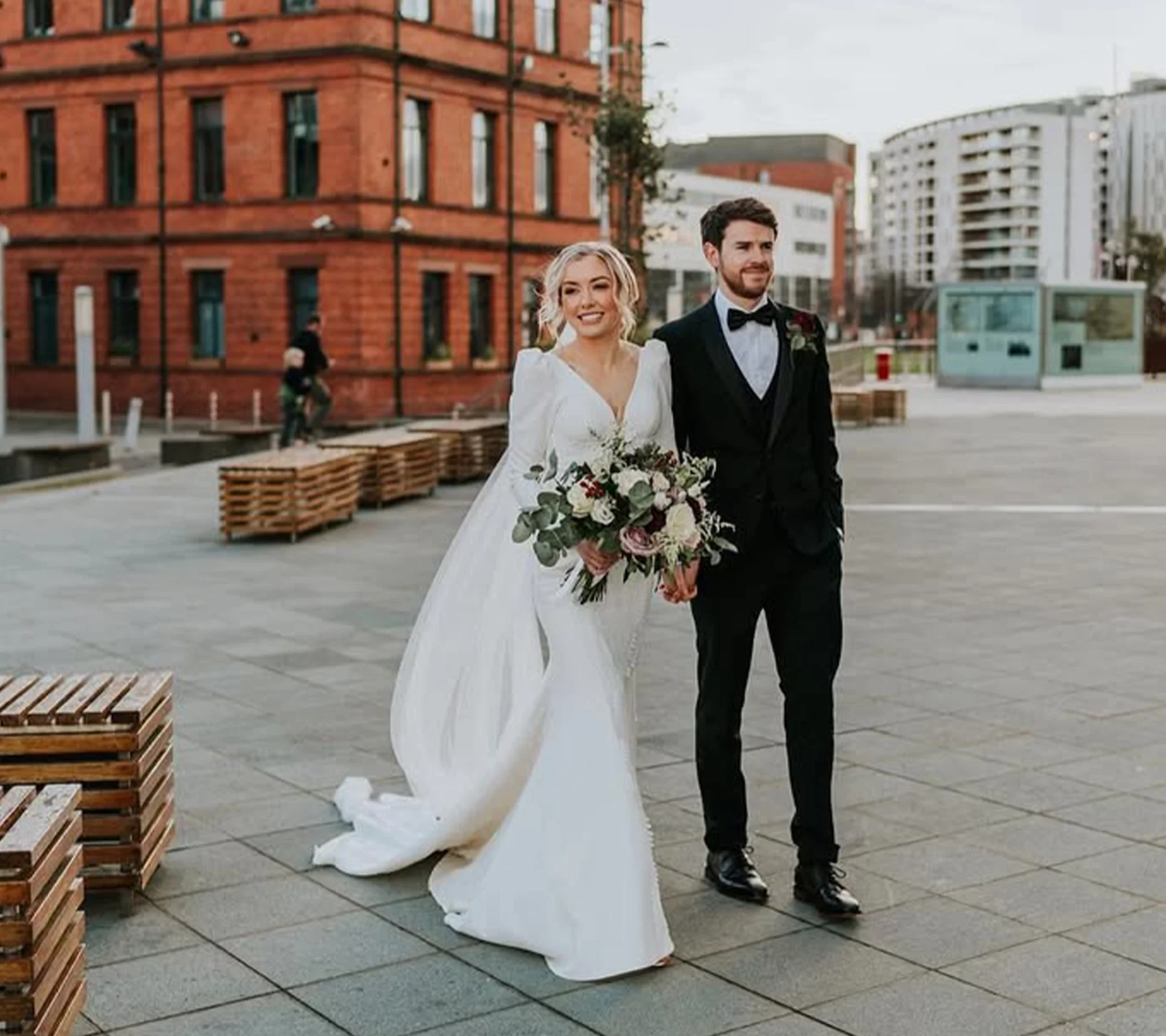 A bride and groom walking together outdoors in an urban setting, with the bride holding a large bouquet of flowers and both smiling.