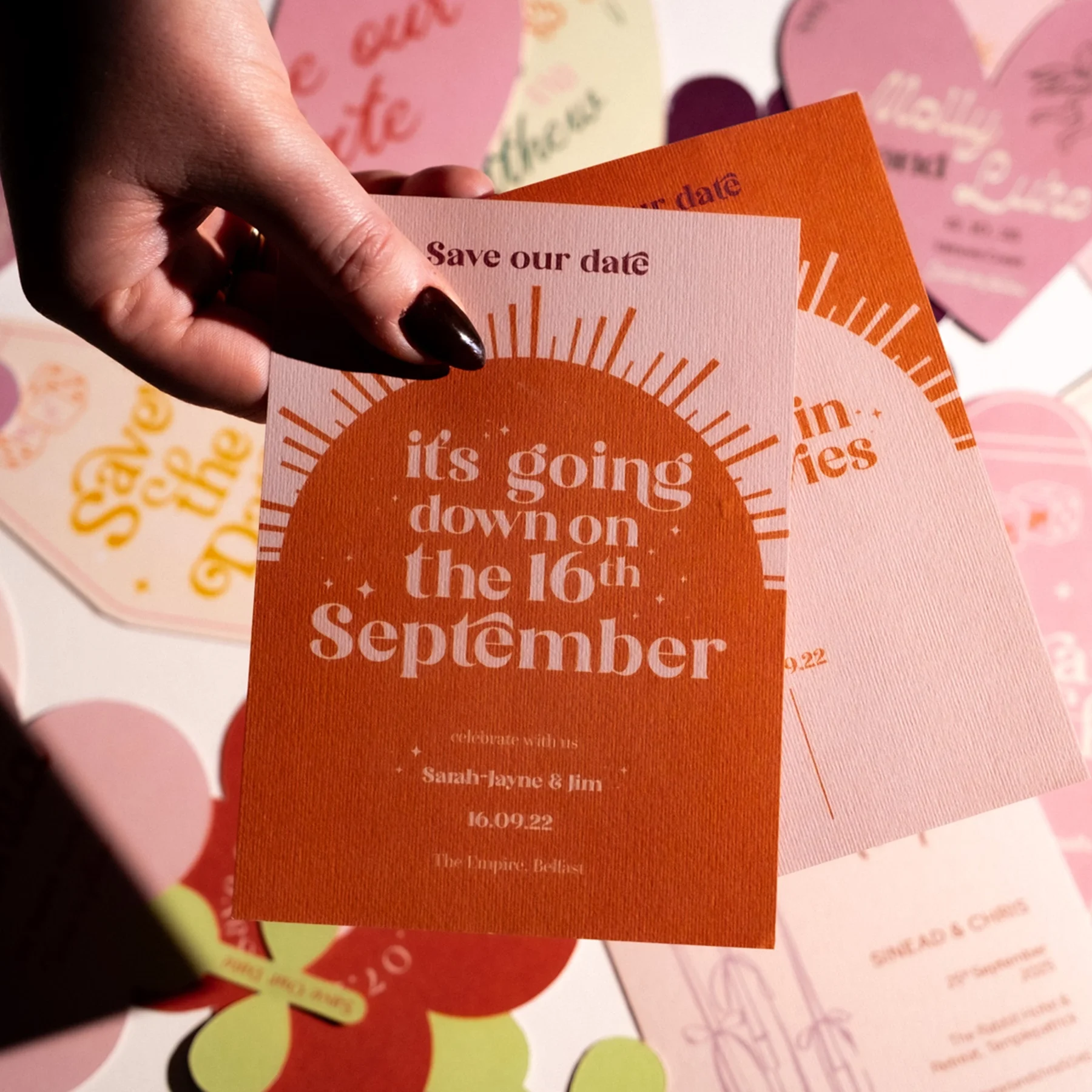 A person's hand holding a wedding save-the-date card with orange and pink colors. The card states the wedding is on September 16th and includes the names Sarah-Jayne and Jim. In the background there are other similar cards and decorations.
