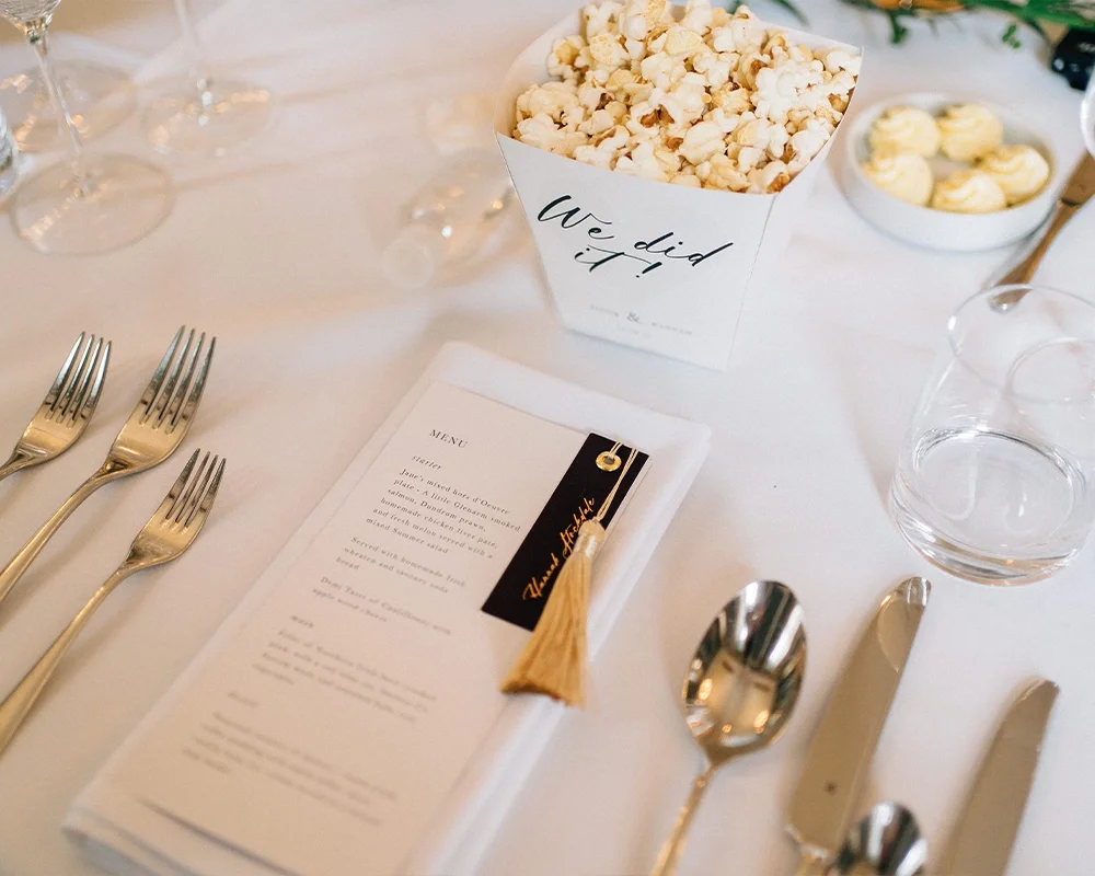 A table setting with multiple forks, a spoon, a butter knife, a drinking glass, and a dinner menu. There is a container of popcorn labeled 'We did it' and a bowl of small desserts or cupcakes.