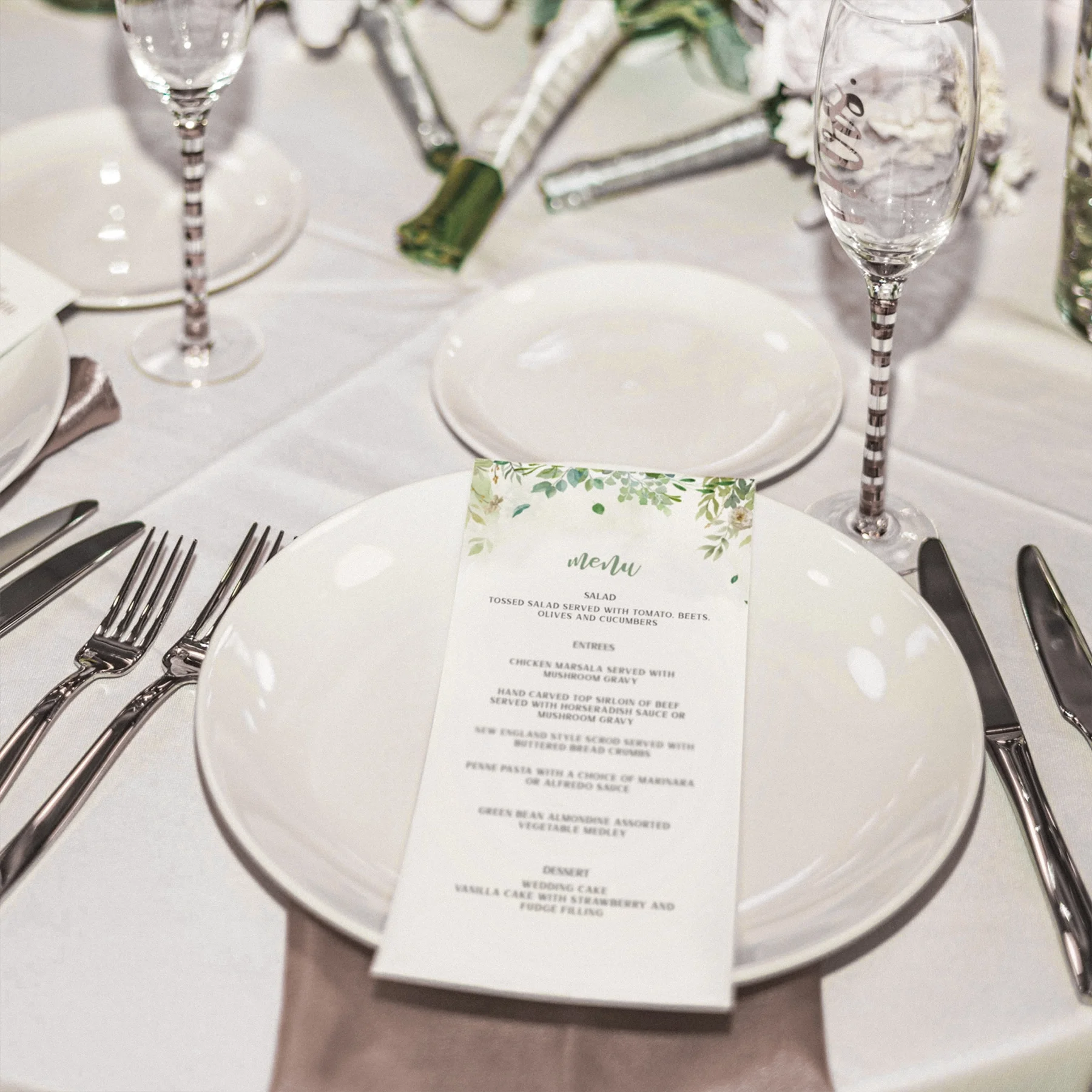 Table setting at a formal event with a white plate holding a printed menu, with silverware on either side, wine glasses, and a floral centerpiece in the background.