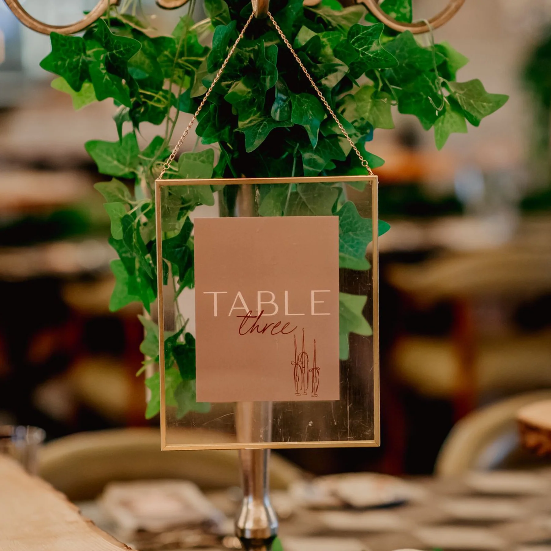 A table card holder hanging from a chain on a plant, with a sign reading 'Table three' and an illustration of three swords.