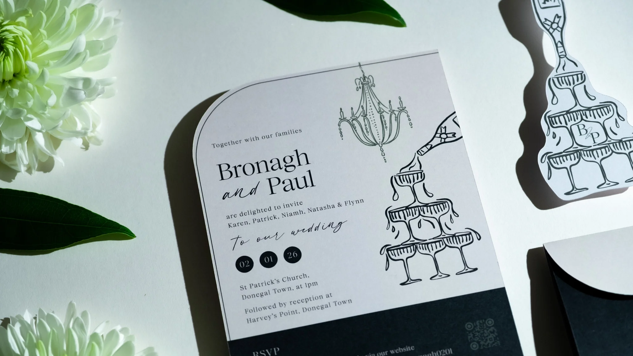 Wedding invitation card for Bronagh and Paul with black line art drawings of champagne glasses and a chandelier, surrounded by white flowers and green leaves.