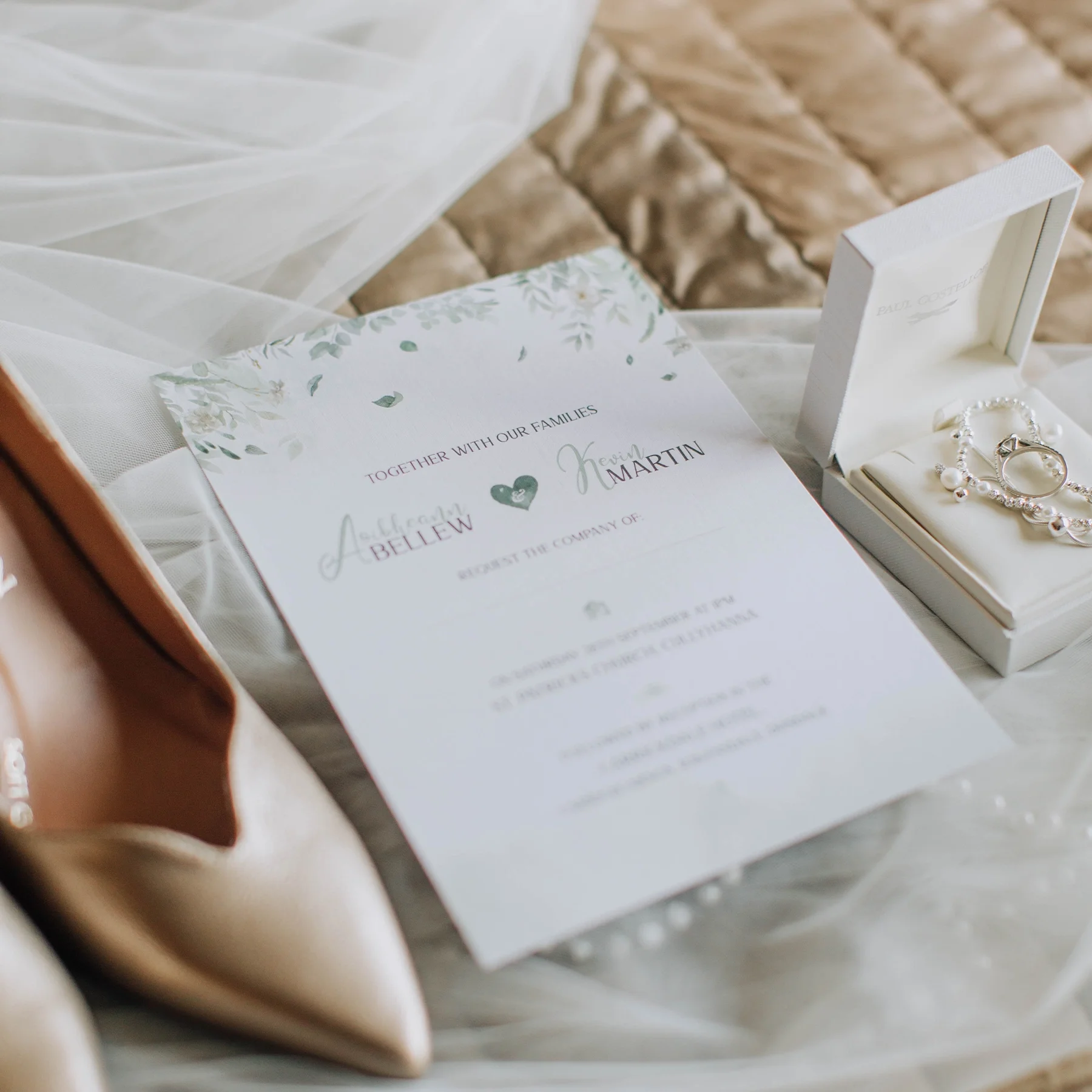 Wedding invitation with floral design, along with jewelry in a white box, a pair of beige high heels, and a veil, all arranged on a soft fabric surface.