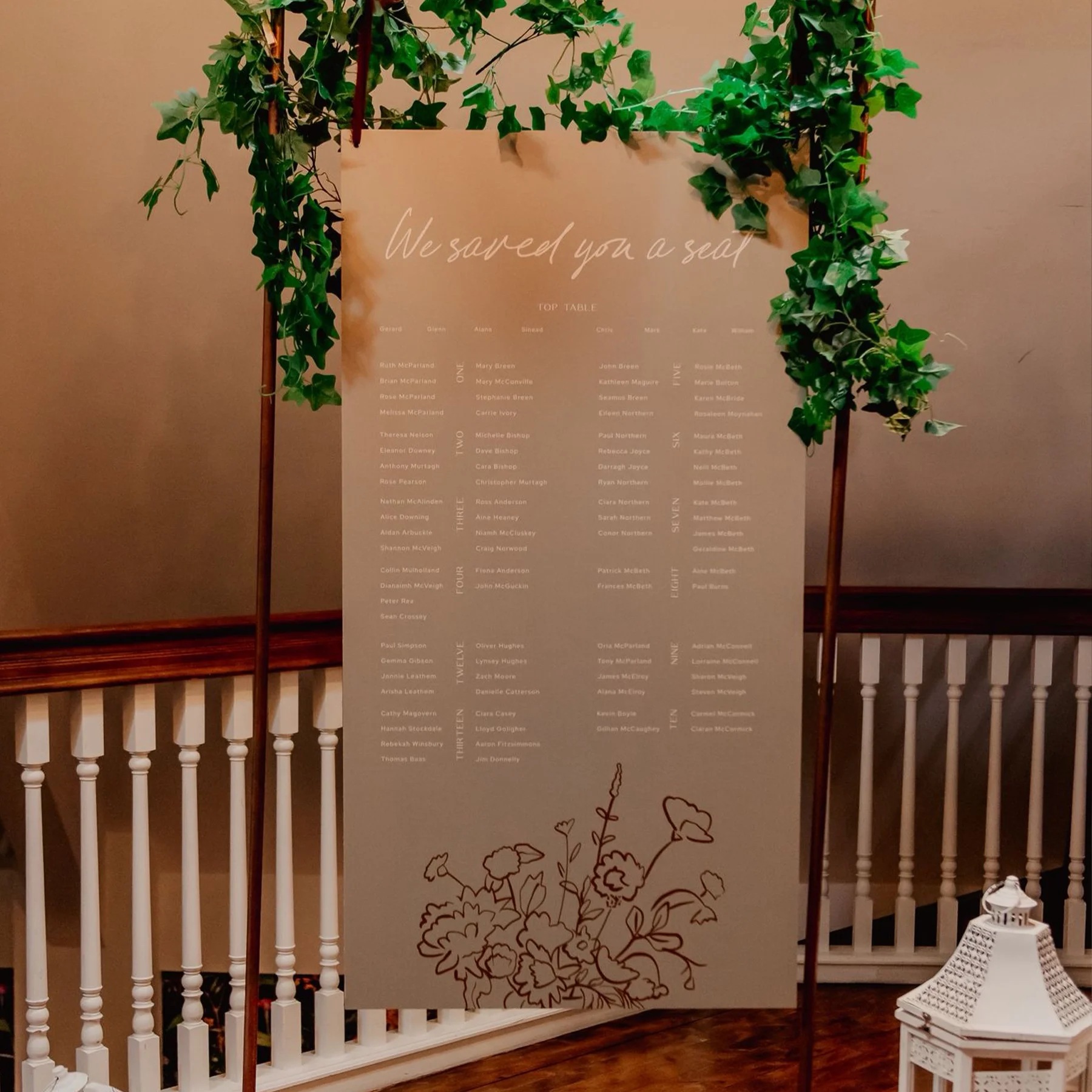 Wedding seating chart sign with the phrase 'We saved you a seat' at the top, decorated with green ivy and floral illustrations at the bottom, displayed on a wooden easel.