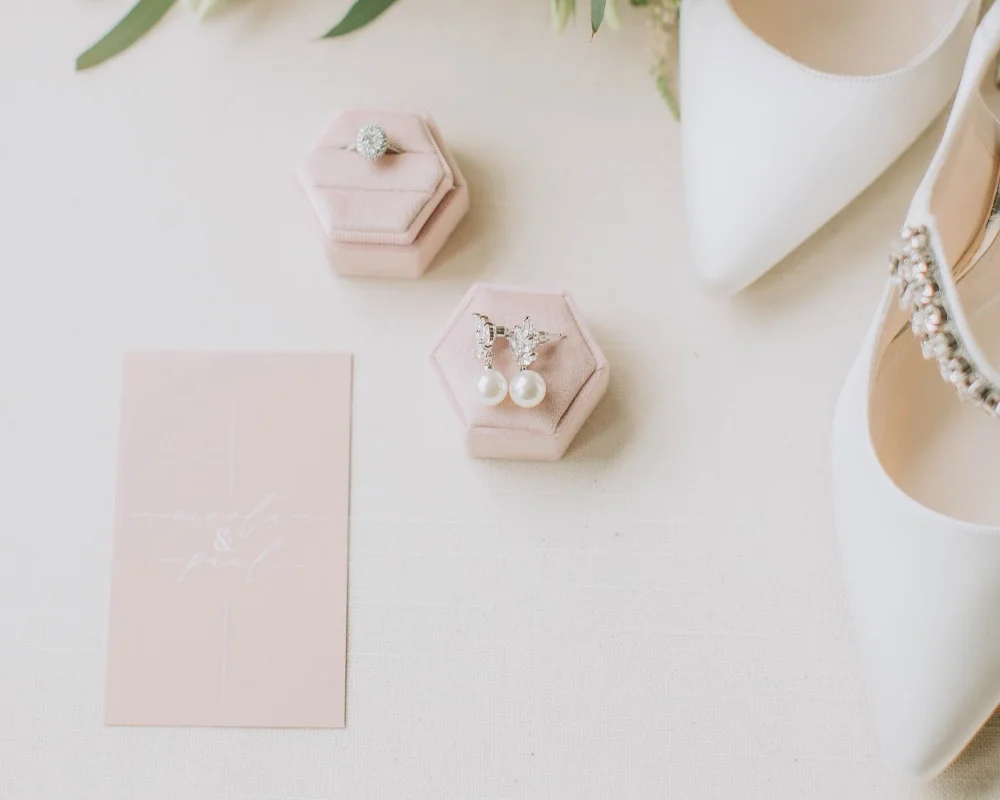 Display of jewelry and wedding accessories including earrings, a ring, and a card on a white surface with heels and a flower in the background.