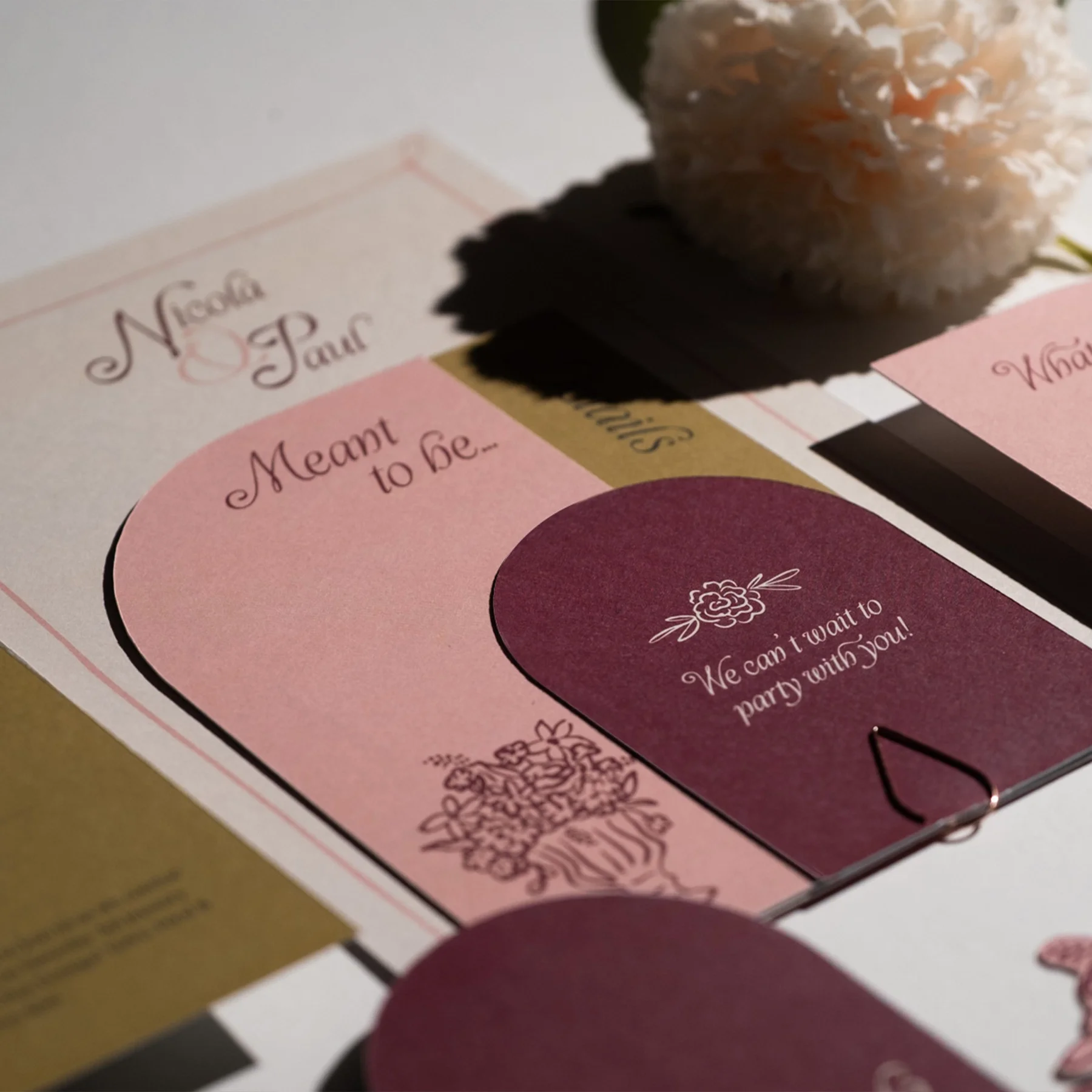 Close-up of wedding or event invitation with pink and purple sections, floral illustrations, and playful fonts, with a white flower in the background.