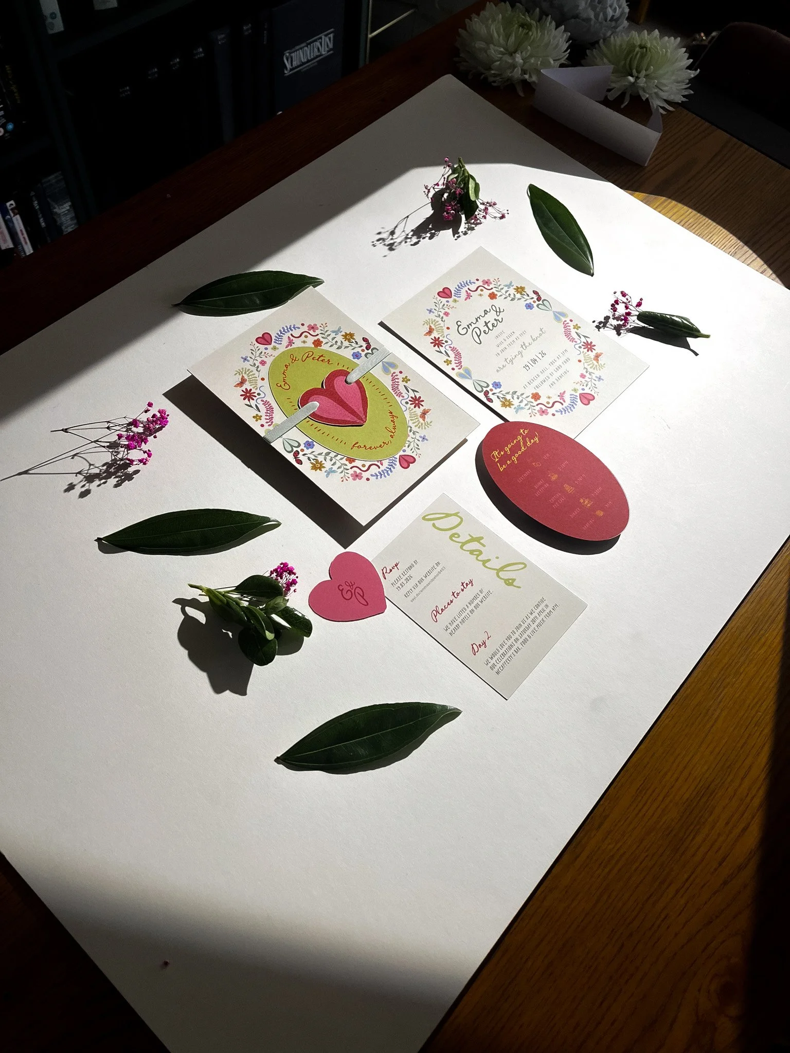 Wedding or event display with floral arrangements and various colorful paper cards, including invitations and details, arranged on a large white sheet on a wooden table.