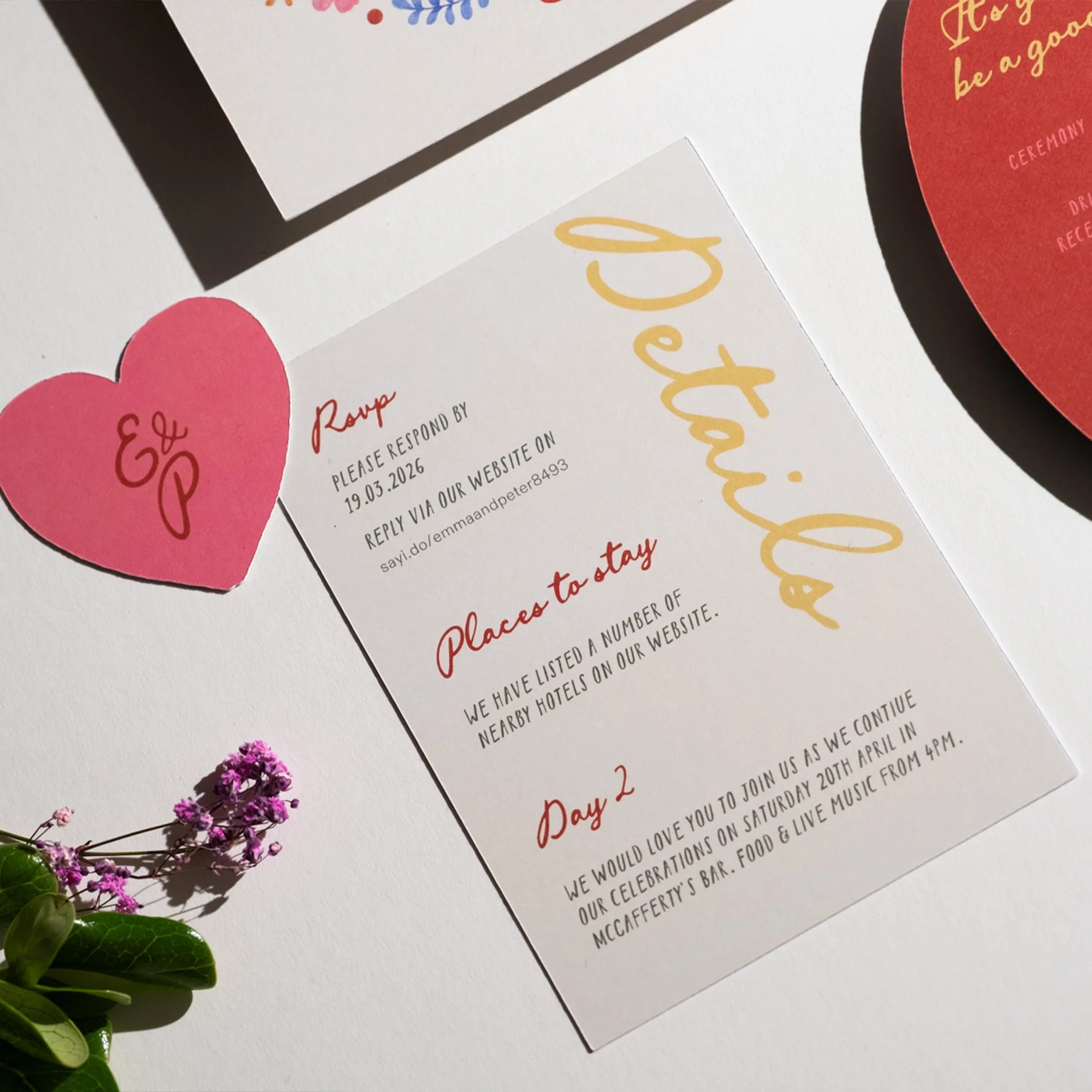 A white event card with yellow and red lettering, includes instructions for a wedding or celebration event, with a pink heart-shaped note with initials 'E & P', and a small purple flower with green leaves.