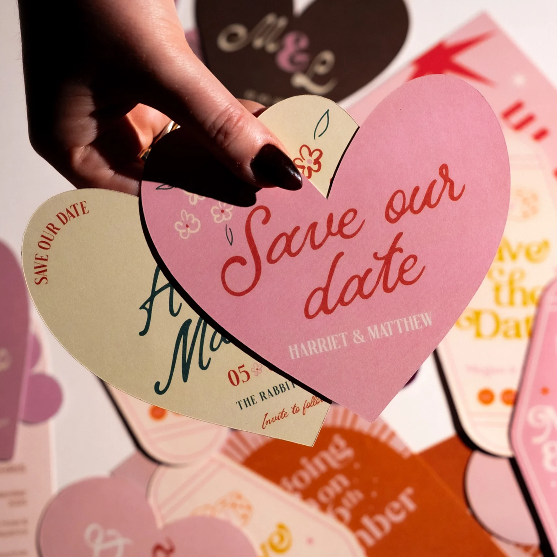 A person holding pink and beige heart-shaped 'Save Our Date' cards with names and wedding details, with other similar cards in the background.