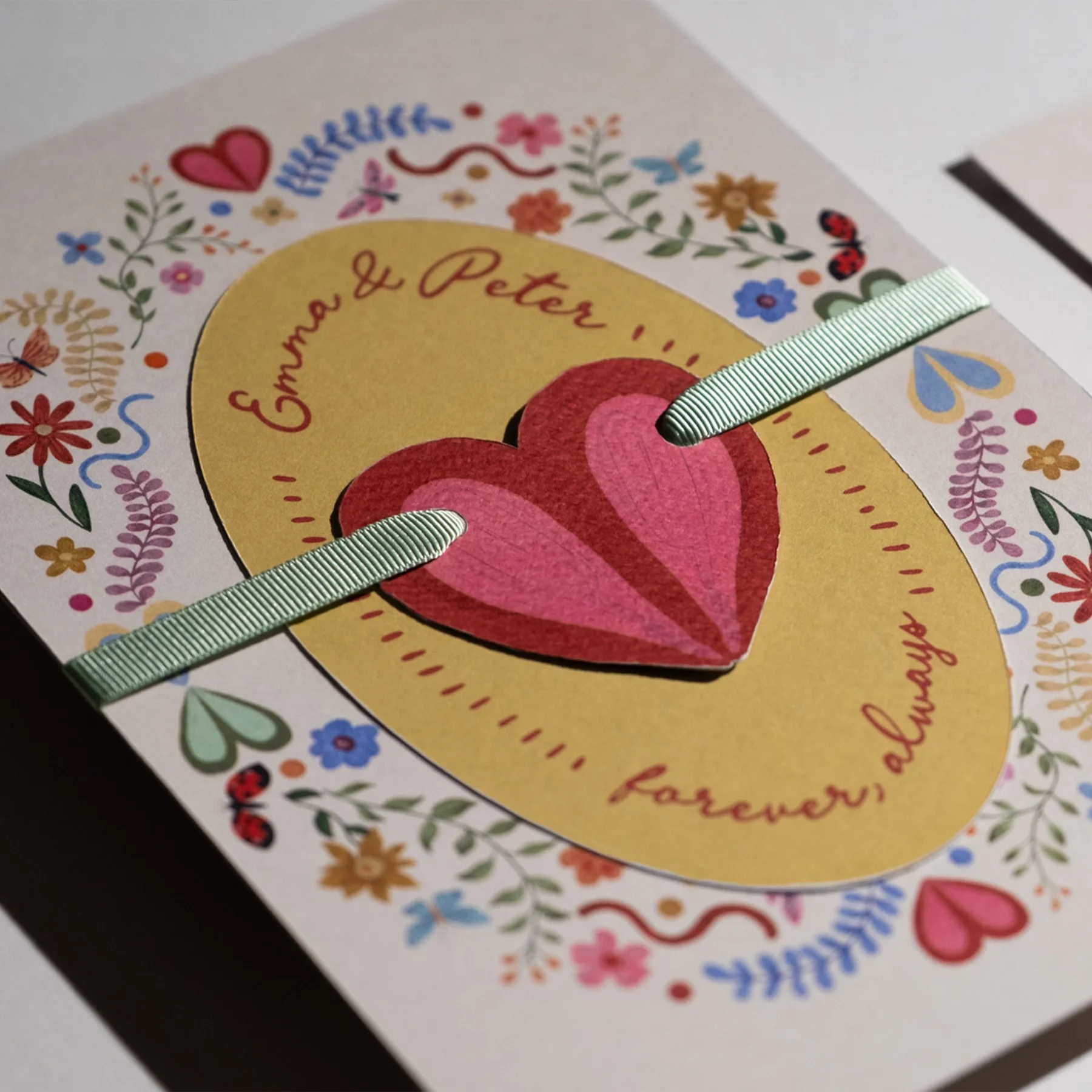 A greeting card with colorful floral and heart decorations. The card has a central illustration of two overlapping pink hearts on a yellow background, with red handwritten text in a foreign language. Two pale green ribbons run horizontally across the card.