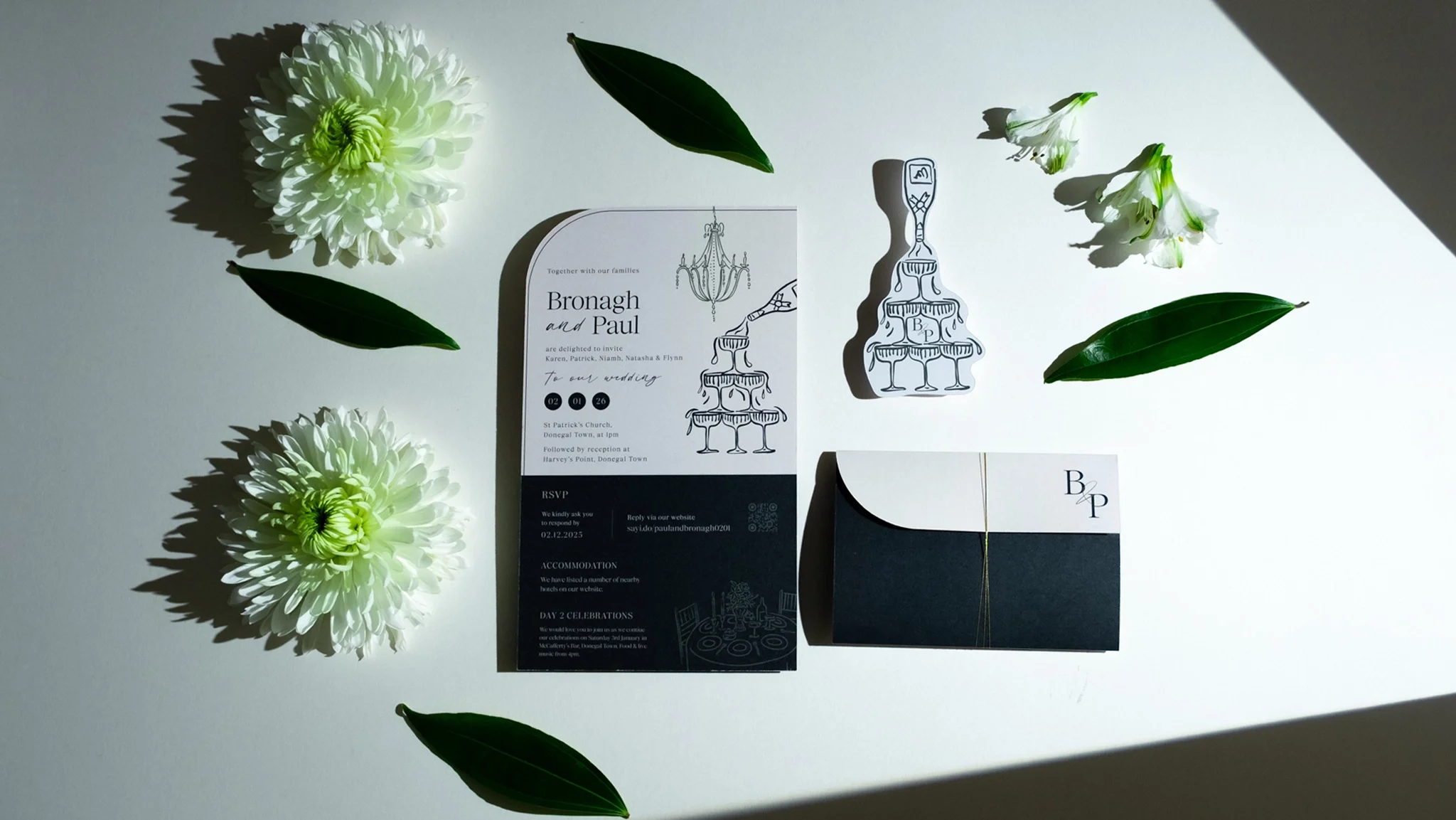 Wedding invitation with white flowers, green leaves, and a small black box on a light surface.