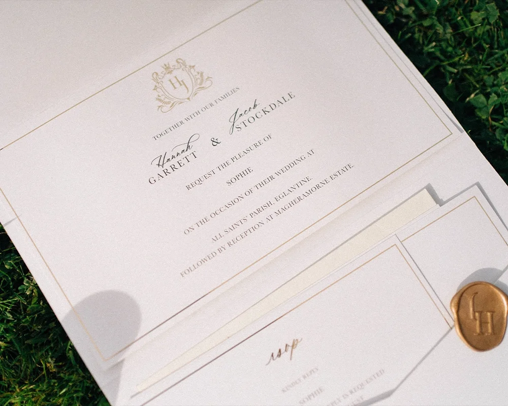A wedding invitation on white paper with gold lettering and a gold wax seal, placed on grass.