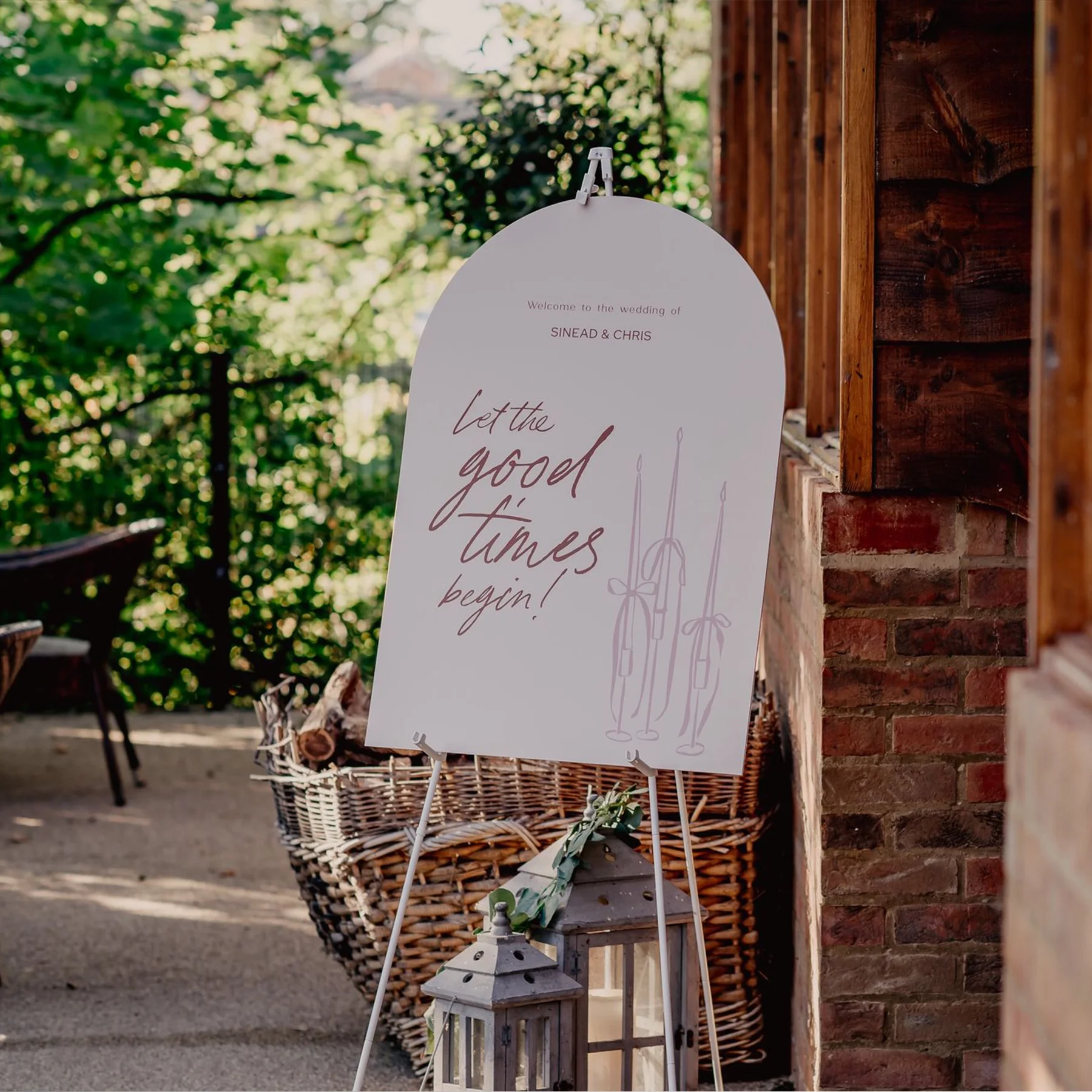 Wedding welcome sign on an easel outdoors with greenery background, decorated with lanterns and a woven basket nearby.