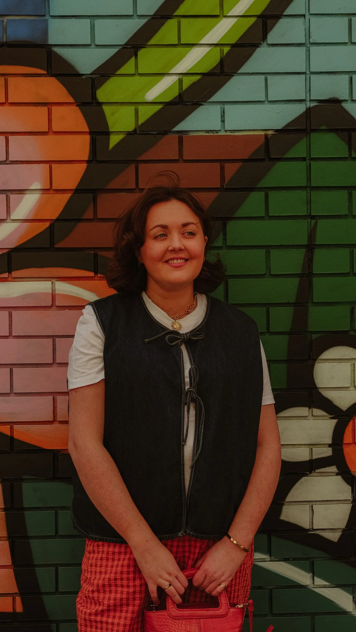 A woman standing in front of a colorful graffiti wall, smiling, holding a pink bag.