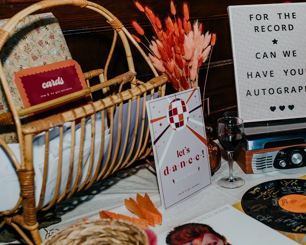 A vintage-style table setup with a wicker basket holding greeting cards, a speech bubble decoration that says "let's dance!" with a disco ball design, a wine glass filled with red wine, a vinyl record, and a whiteboard with black letters asking "For 