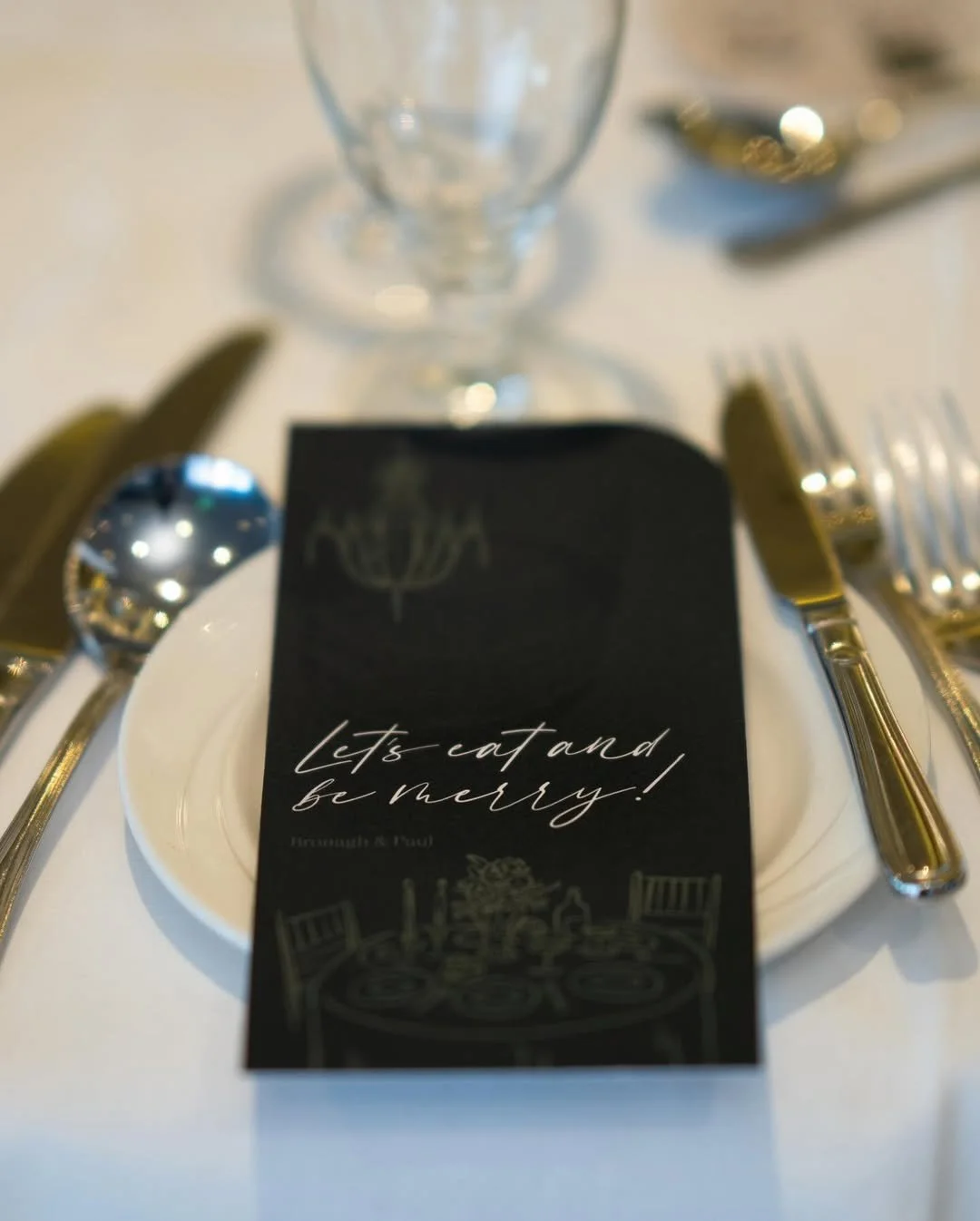 A table setting with a black card that has the message 'Let's eat and be merry!' placed on a white plate, surrounded by silverware, a glass, and a blurred background.
