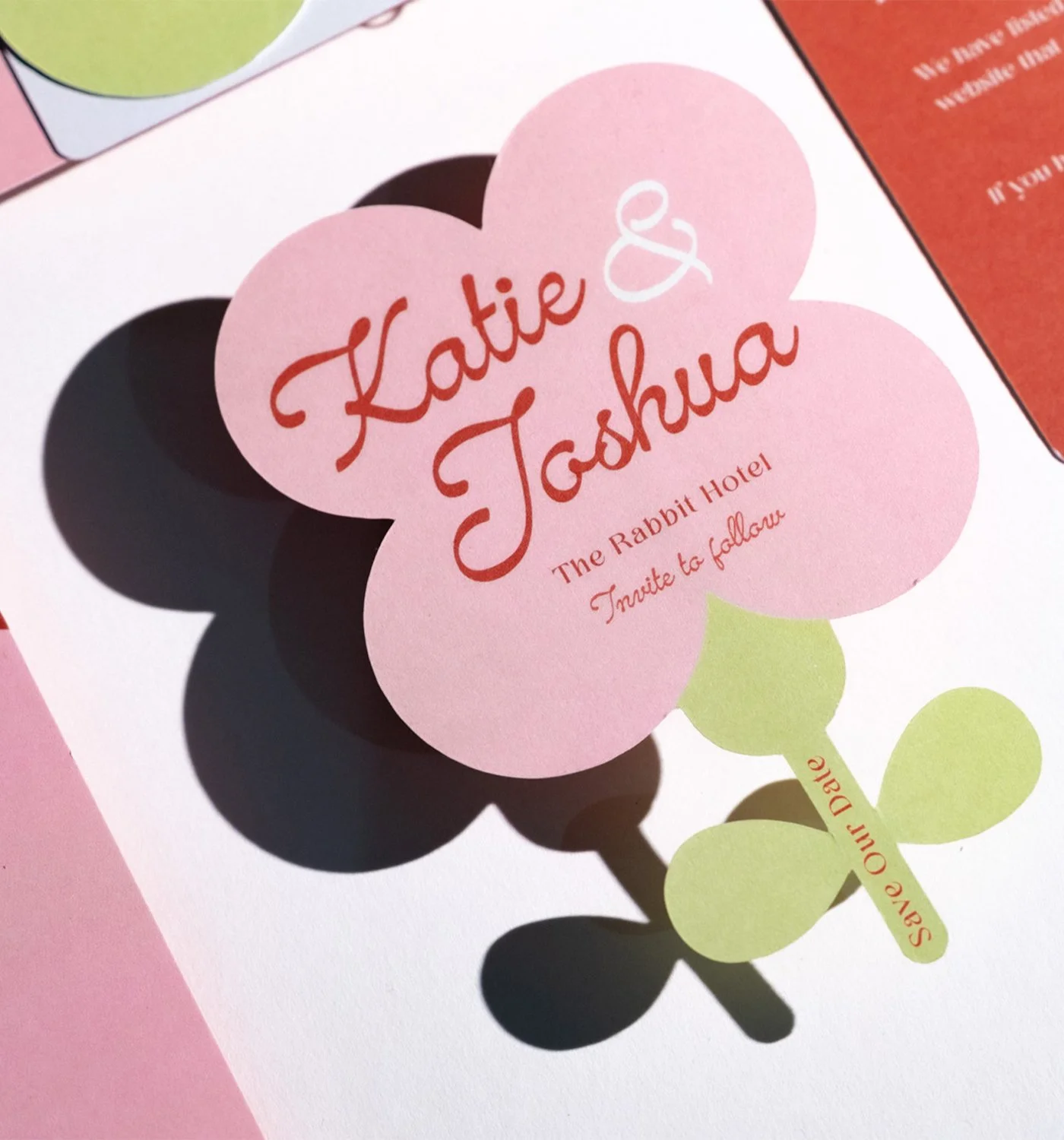 Pink flower design with red cursive text reading 'Katie & Joshua' and smaller red text 'The Rabbit Hotel' and 'Invite to follow'. Green leaves with red text 'Save Our Date' beneath the flower.