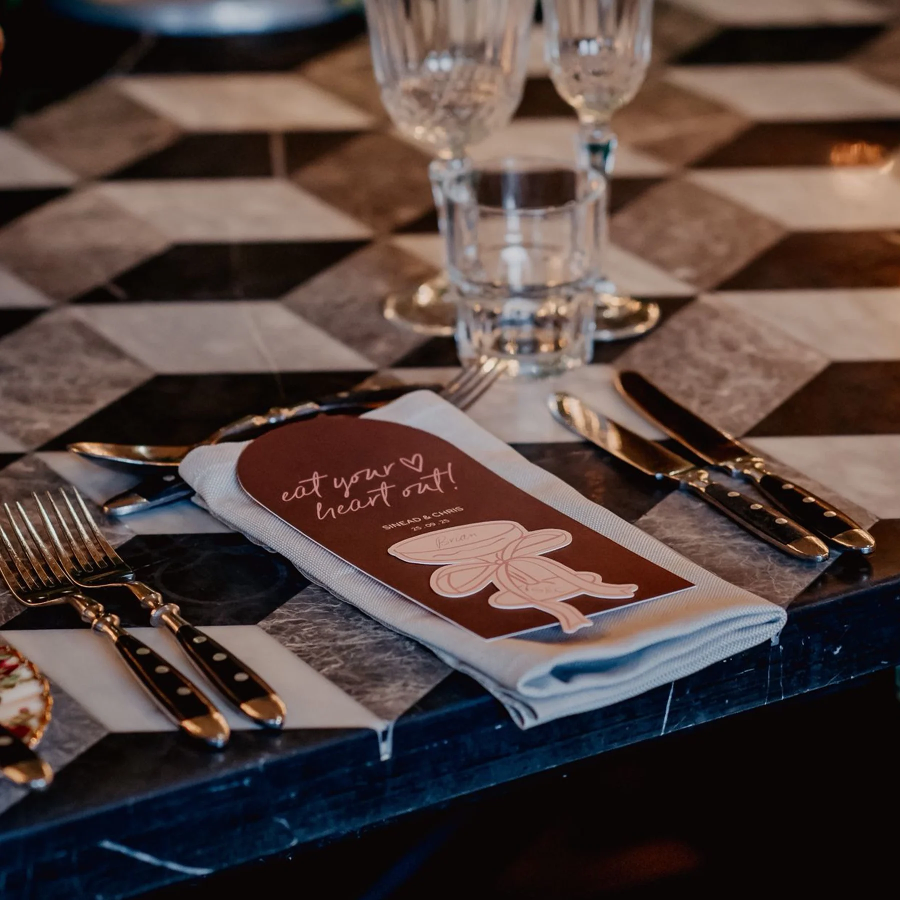 Table setting with a white napkin, silverware including a fork, knife, and spoon, a water glass, and two champagne glasses on a patterned black and white tablecloth. A paper menu or card with the text 'eat your heart out!' and a heart is placed on th