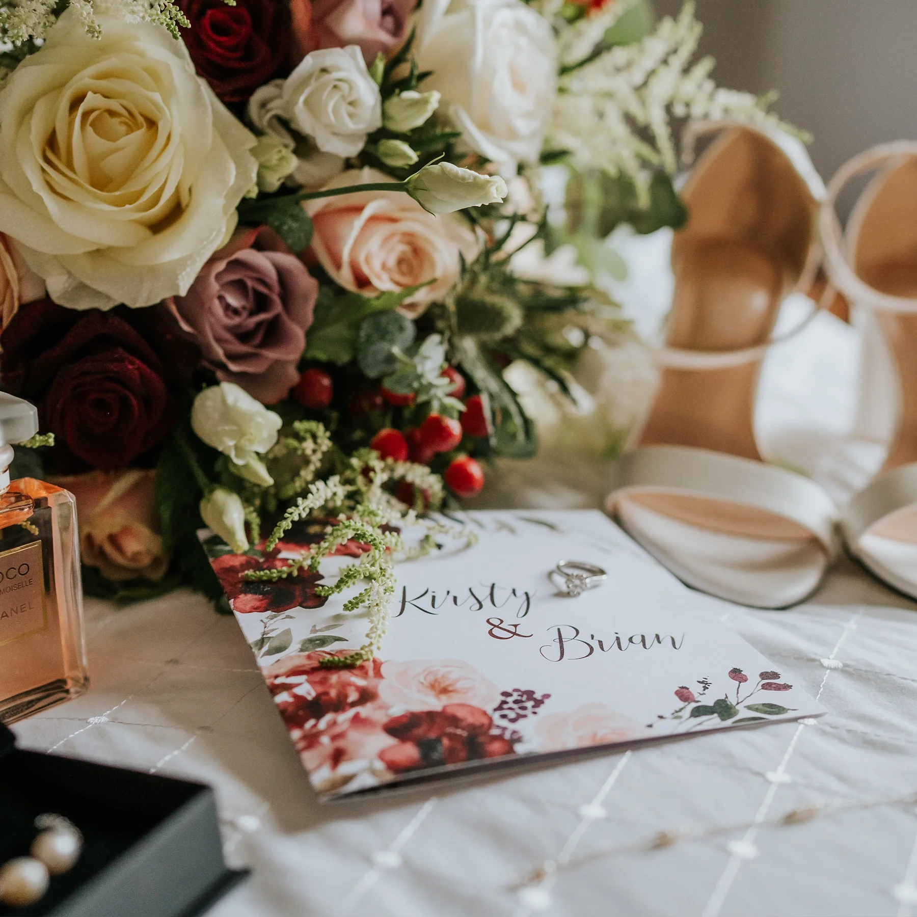 A wedding or event table arrangement with a floral centerpiece, a card with the names Kirsty and Brian, a ring, a perfume bottle, and a pair of nude-colored heels.